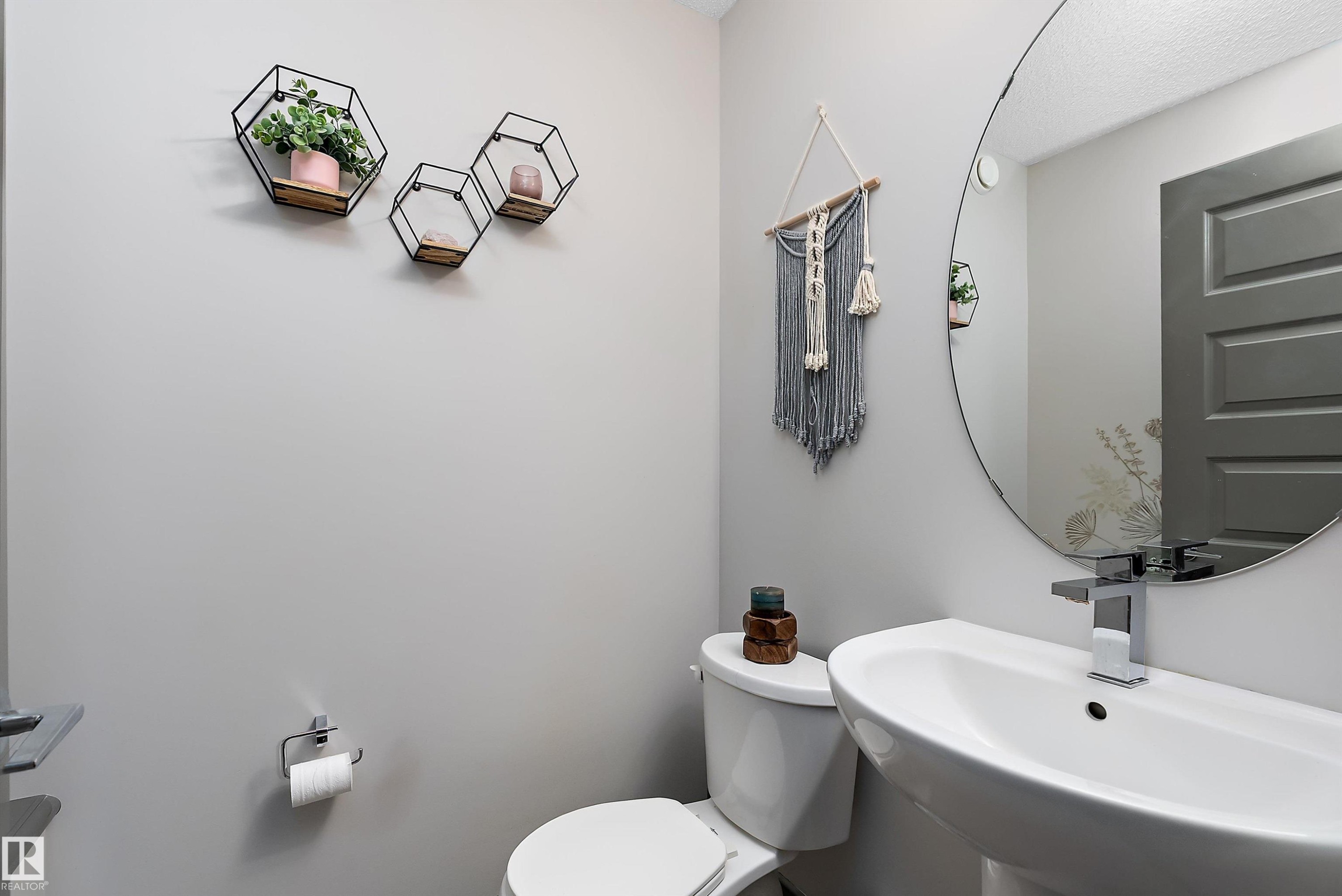 The bathroom features a white pedestal sink with a chrome faucet, a round wall-mounted mirror, and light gray walls - 7776 Eifert Crescent Nw, Edmonton, AB - Indoor Photo Showing Bathroom
