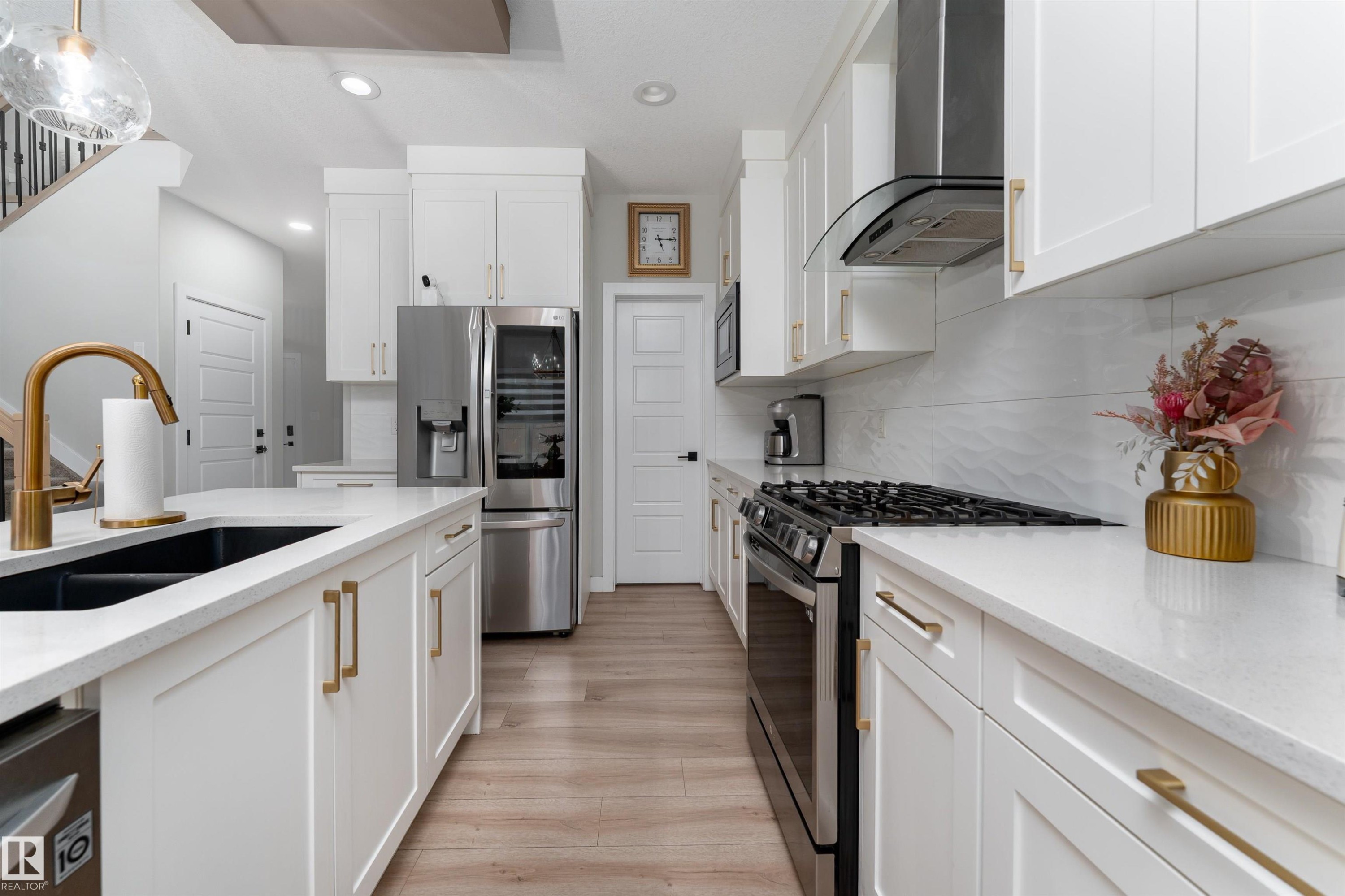 The kitchen features white cabinetry with gold-toned hardware, white countertops, and stainless steel appliances including a refrigerator and range with a hood - 993 Berg Pl, Leduc, AB - Indoor Photo Showing Kitchen With Double Sink With Upgraded Kitchen