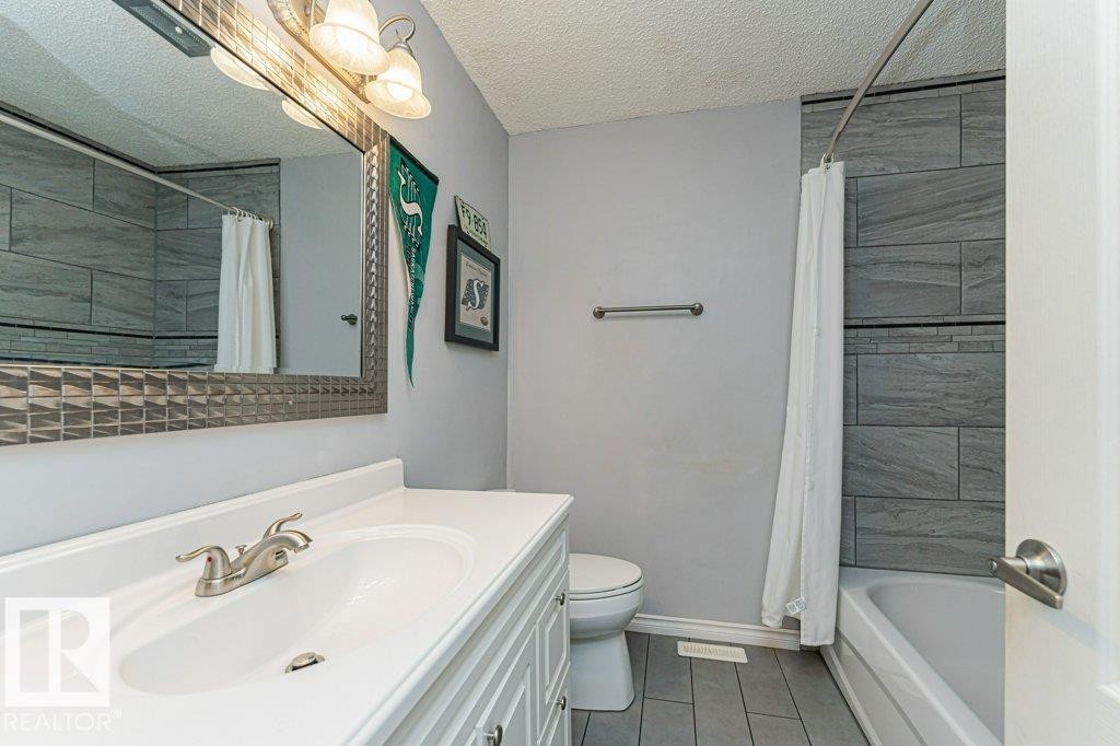 The bathroom features a vanity with an integrated sink and brushed nickel faucet, a mirror with an ornate frame and overhead lighting, a toilet, and a shower-tub combination with grey tile surround and a shower curtain - 8 Duke Drive, Lamont, AB - Indoor Photo Showing Bathroom
