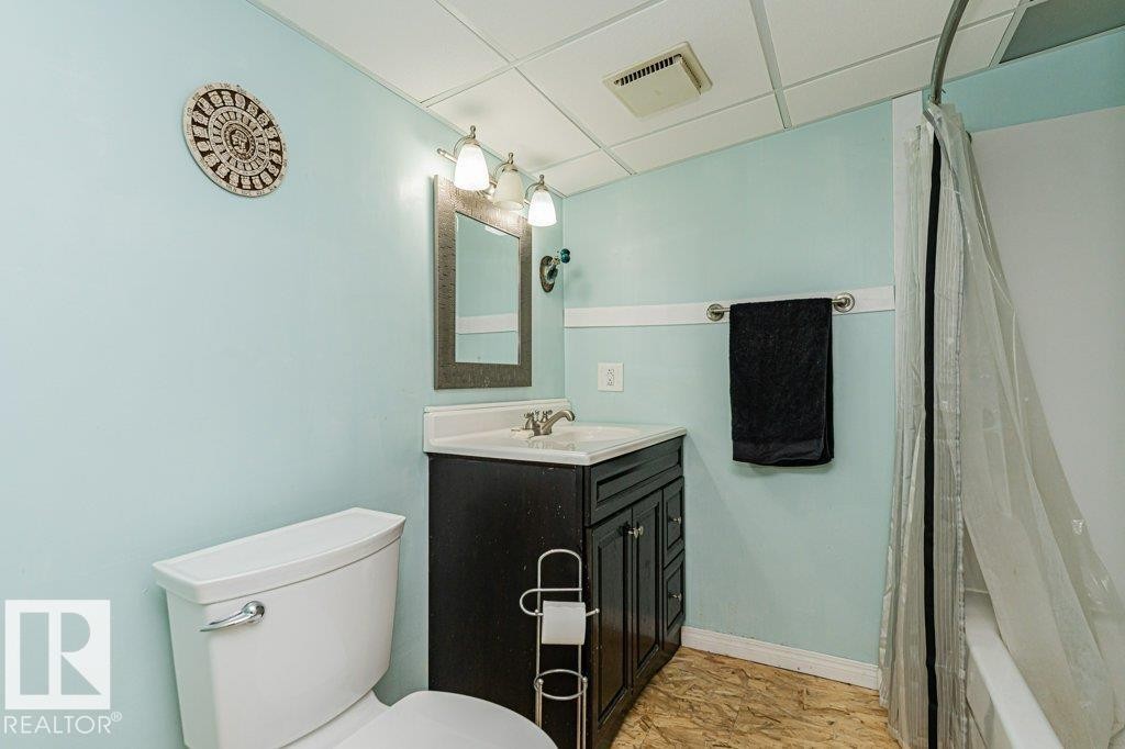 The bathroom features a toilet, a vanity with a mirror and overhead lighting, and a shower with a clear shower curtain - 8 Duke Drive, Lamont, AB - Indoor Photo Showing Bathroom