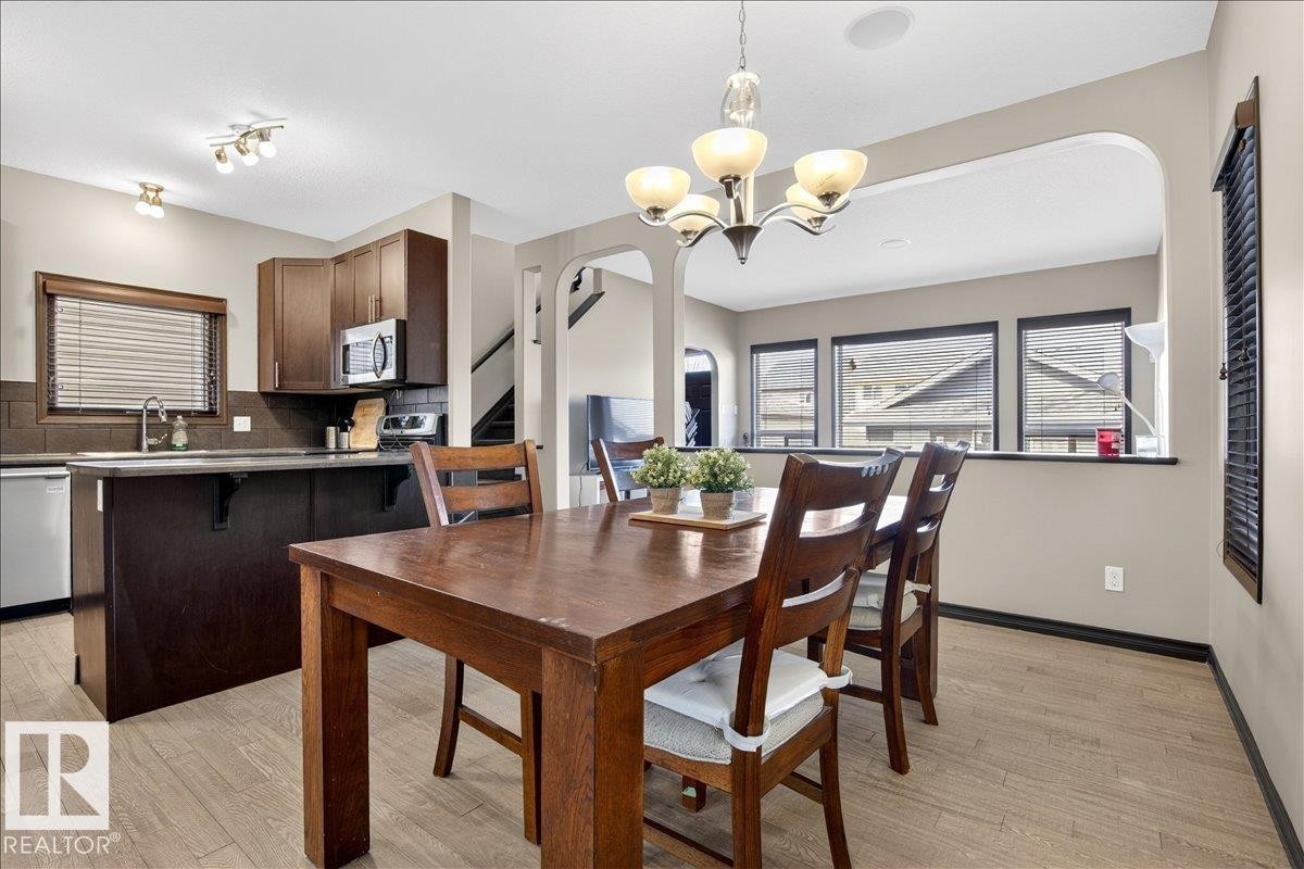 Dining area with a chandelier and light-toned flooring, open to a kitchen featuring dark wood cabinetry, stainless steel appliances, and a window with blinds - 1530 Chapman Way, Edmonton, AB - Indoor