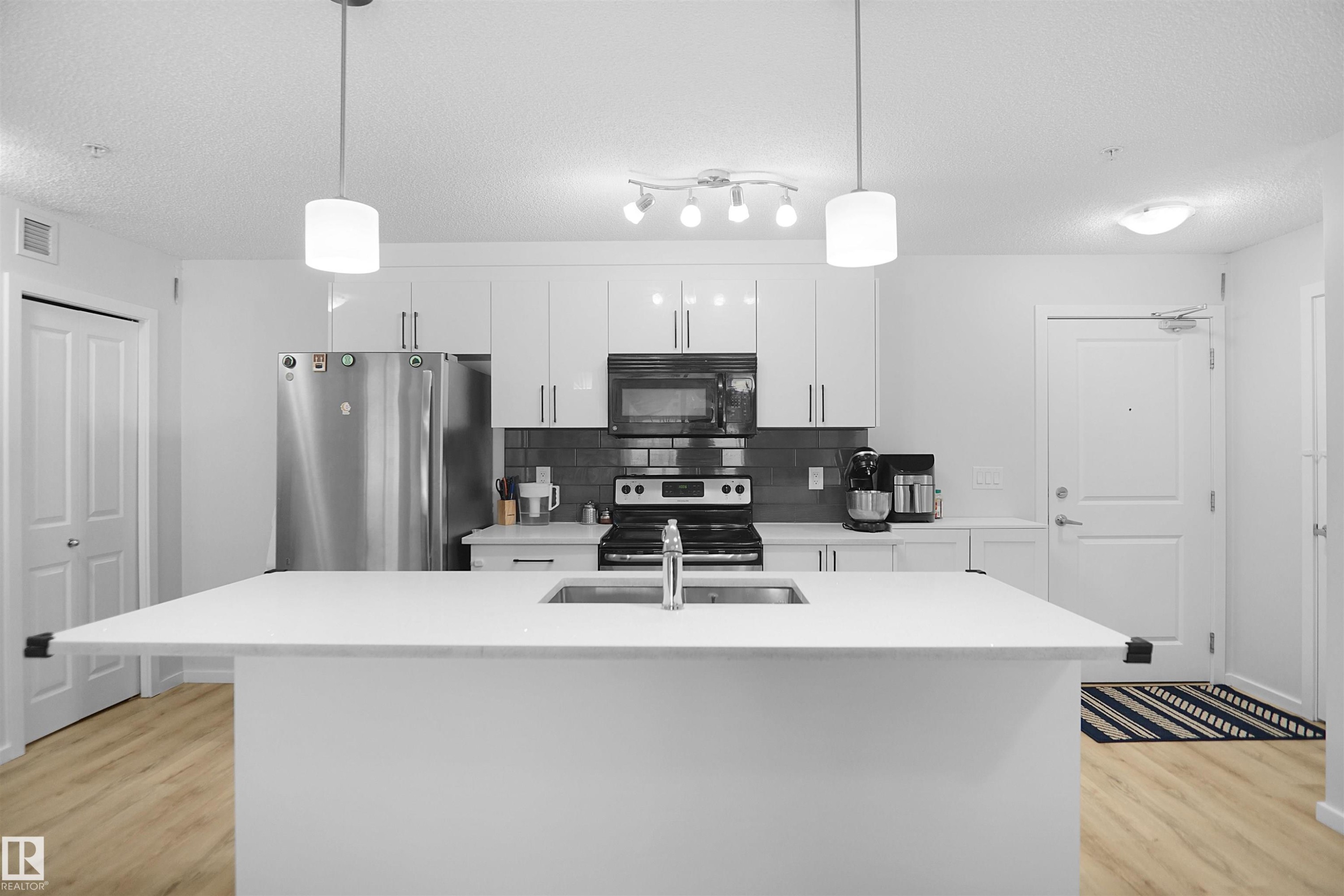 The kitchen features a spacious island with an integrated sink, white cabinetry, a dark tiled backsplash, and stainless steel appliances - 103 3353 16A Avenue, Edmonton, AB - Indoor Photo Showing Kitchen With Double Sink With Upgraded Kitchen
