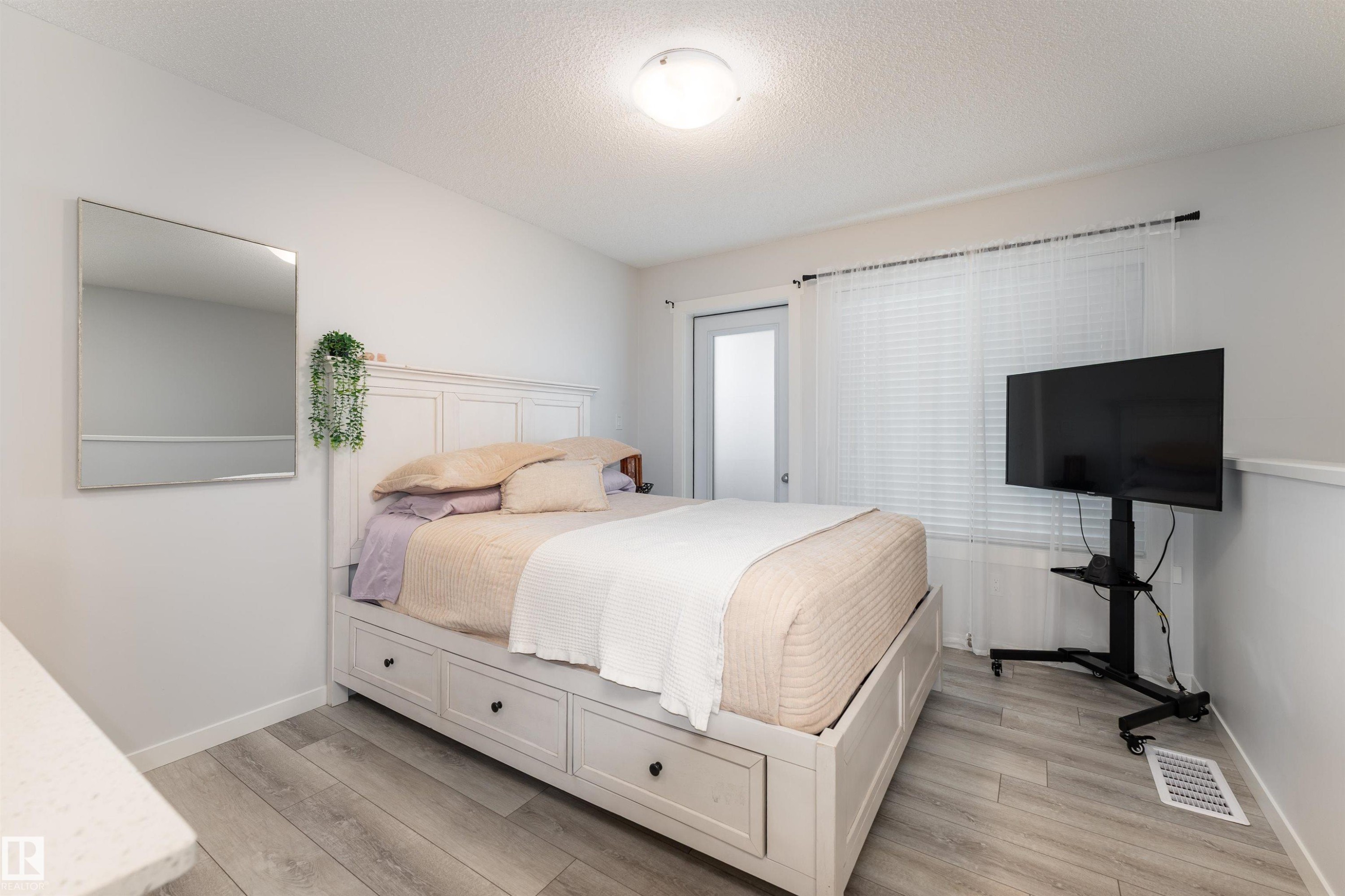 This room features light-toned flooring, a light-colored headboard with panel detailing, and a door leading to an exterior space - 6 12815 Cumberland Rd, Edmonton, AB - Indoor Photo Showing Bedroom