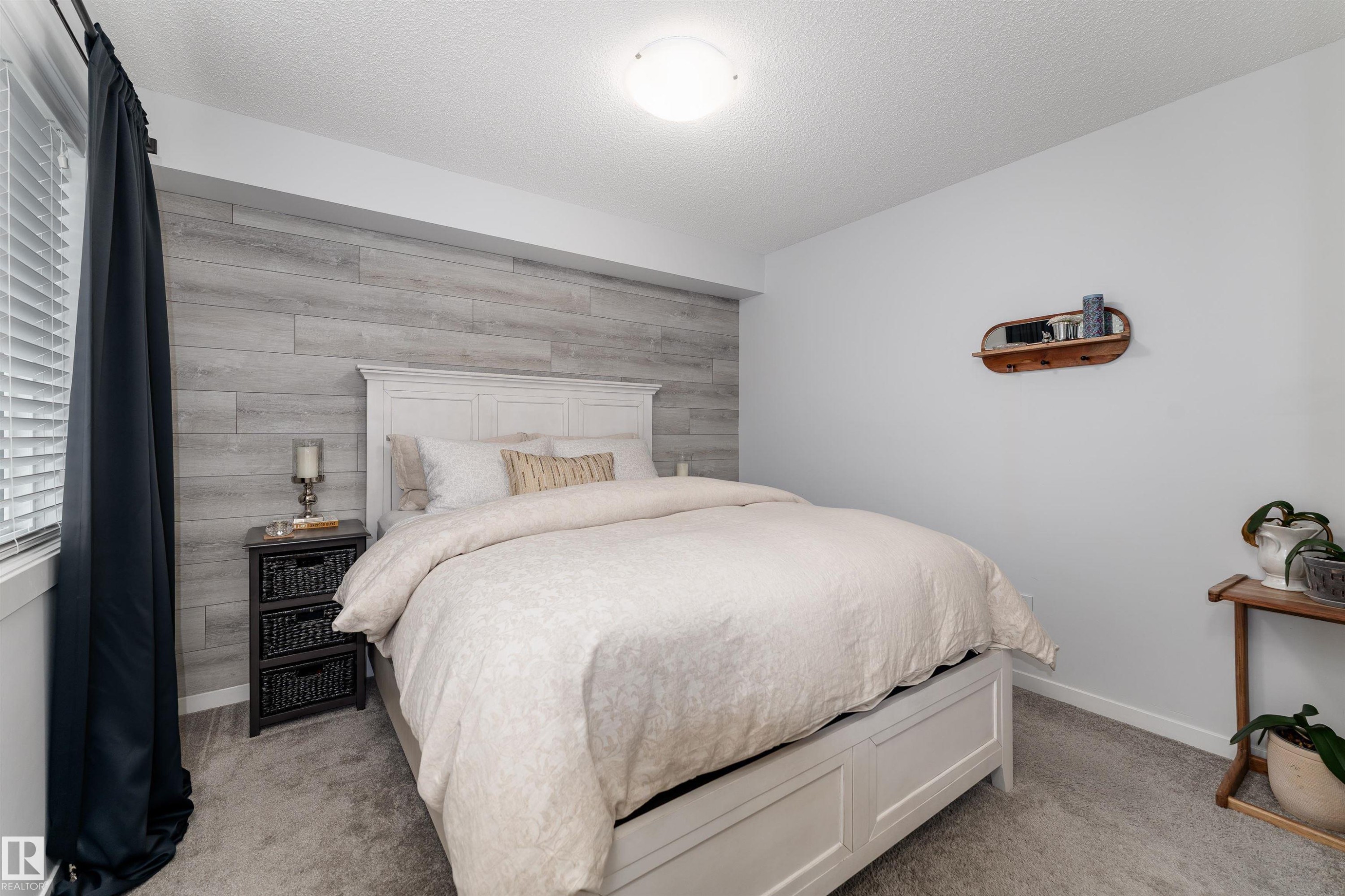 This inviting room features a light grey textured accent wall, a window with blinds and dark curtains, and light grey carpeting - 6 12815 Cumberland Rd, Edmonton, AB - Indoor Photo Showing Bedroom