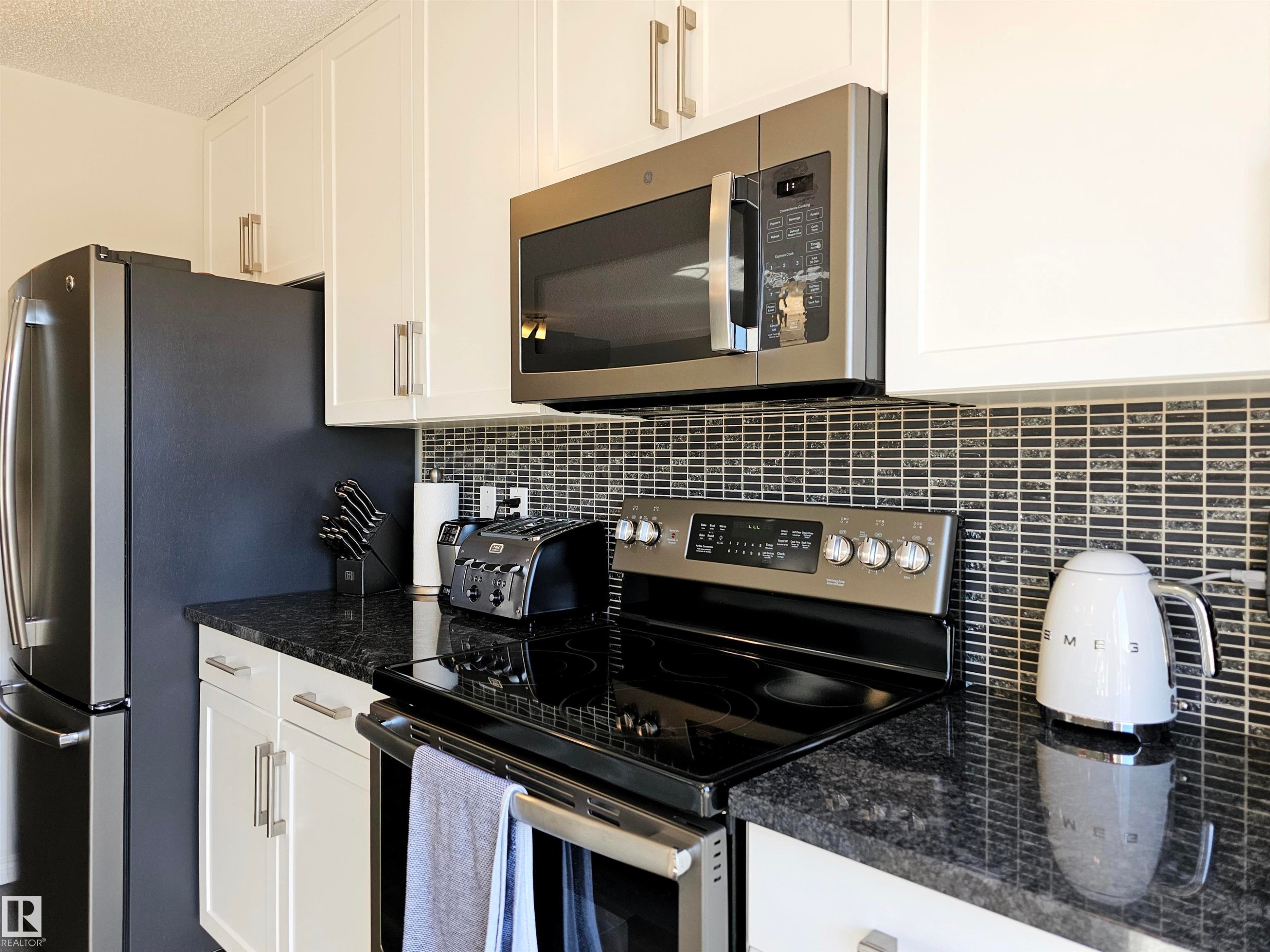 The kitchen features white cabinetry, dark countertops, a tile backsplash, and stainless steel appliances including a refrigerator, microwave, and range - 6888 Evans Wynd, Edmonton, AB - Indoor Photo Showing Kitchen With Stainless Steel Kitchen With Upgraded Kitchen