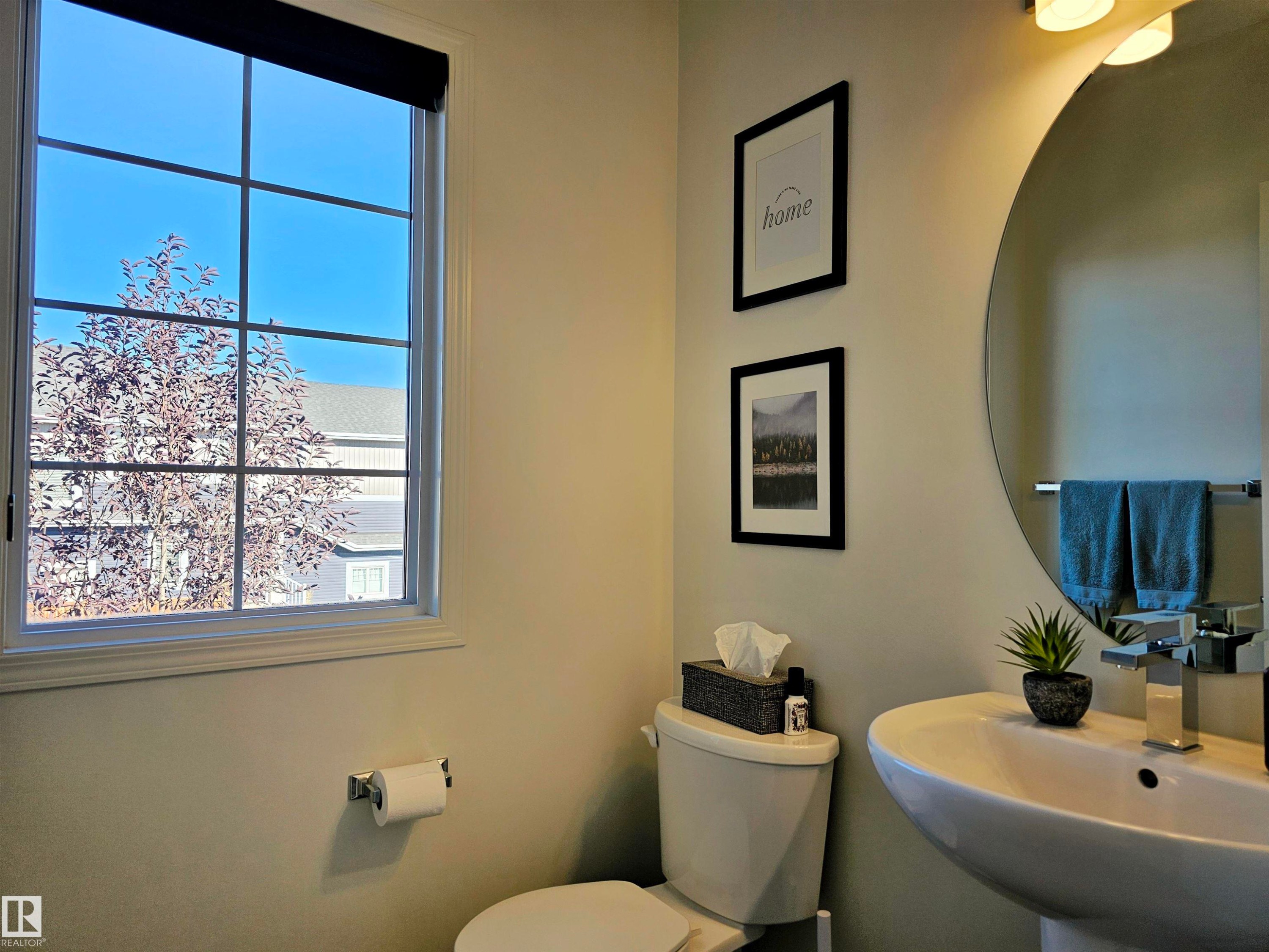 A well-appointed powder room featuring a window with a black roller blind, a pedestal sink with a chrome faucet, and a large round mirror - 6888 Evans Wynd, Edmonton, AB - Indoor Photo Showing Bathroom