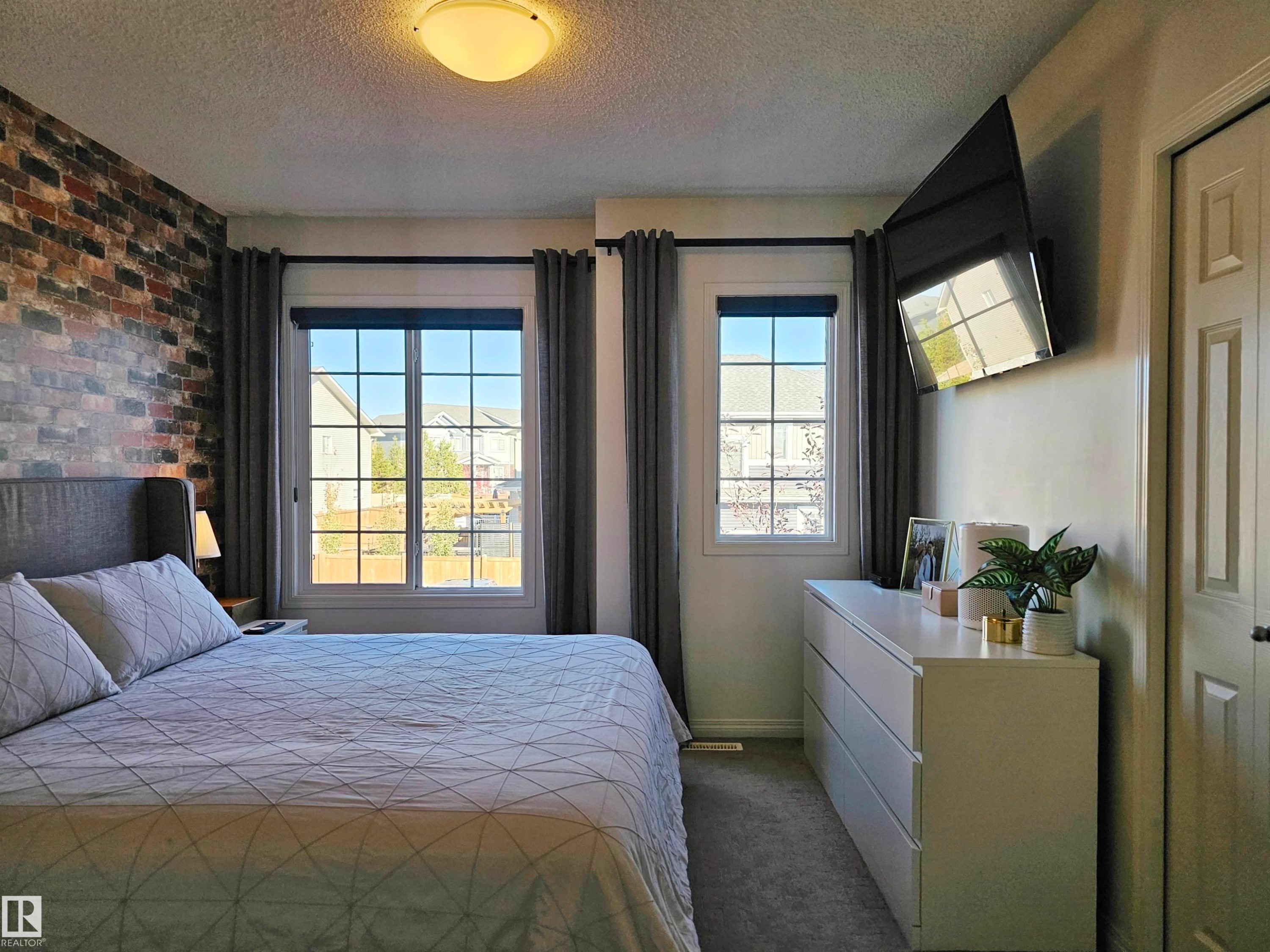 This space features two windows with dark grey curtains, a brick accent wall, and a light-colored ceiling fixture - 6888 Evans Wynd, Edmonton, AB - Indoor Photo Showing Bedroom