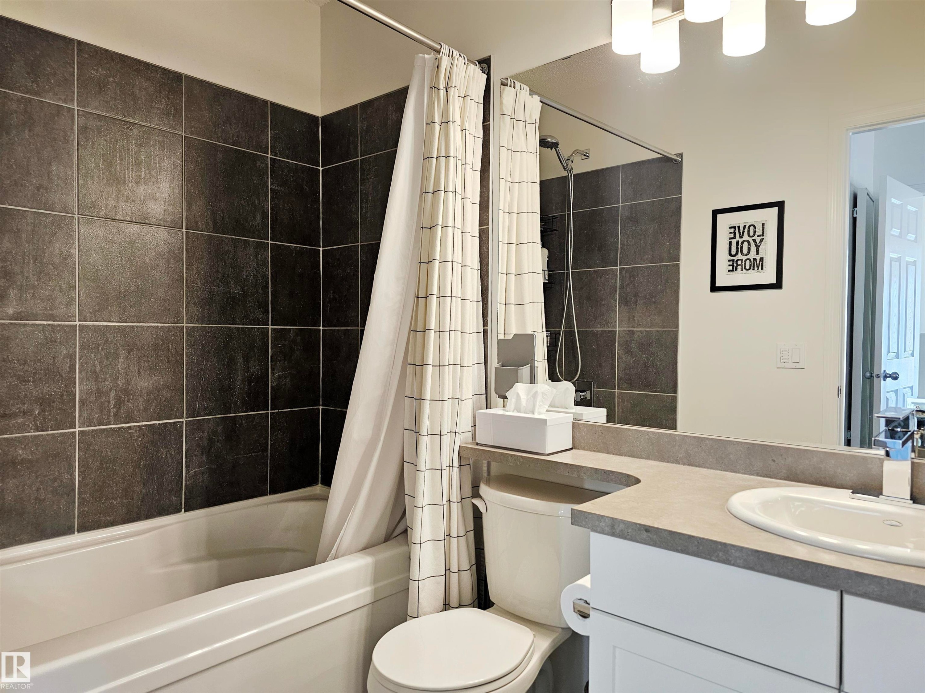 Bathroom featuring a bathtub with dark tiled surround, a toilet, a vanity with a white countertop, and a mirror with overhead lighting - 6888 Evans Wynd, Edmonton, AB - Indoor Photo Showing Bathroom