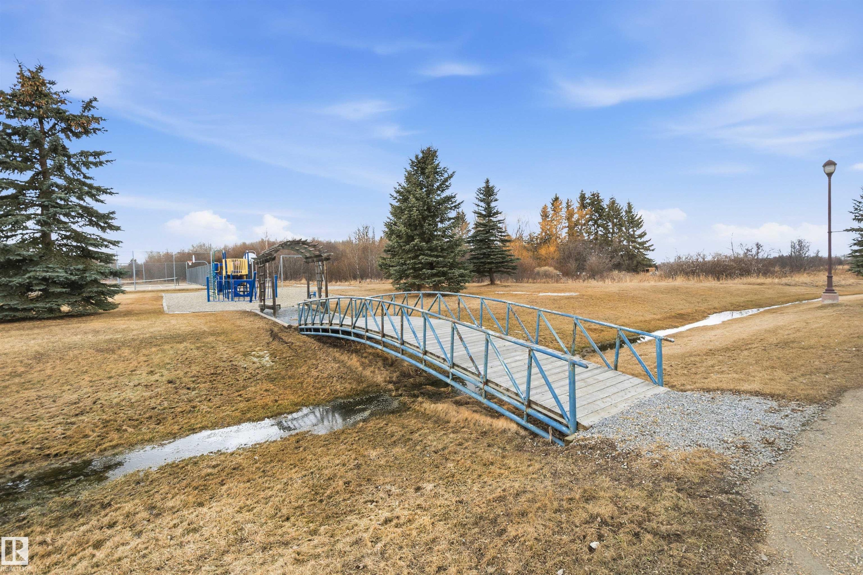 A picturesque outdoor area featuring a bridge over a small stream, a playground, and mature trees, all under a clear sky - 17 Sunset Harbour, Rural Wetaskiwin County, AB - Outdoor