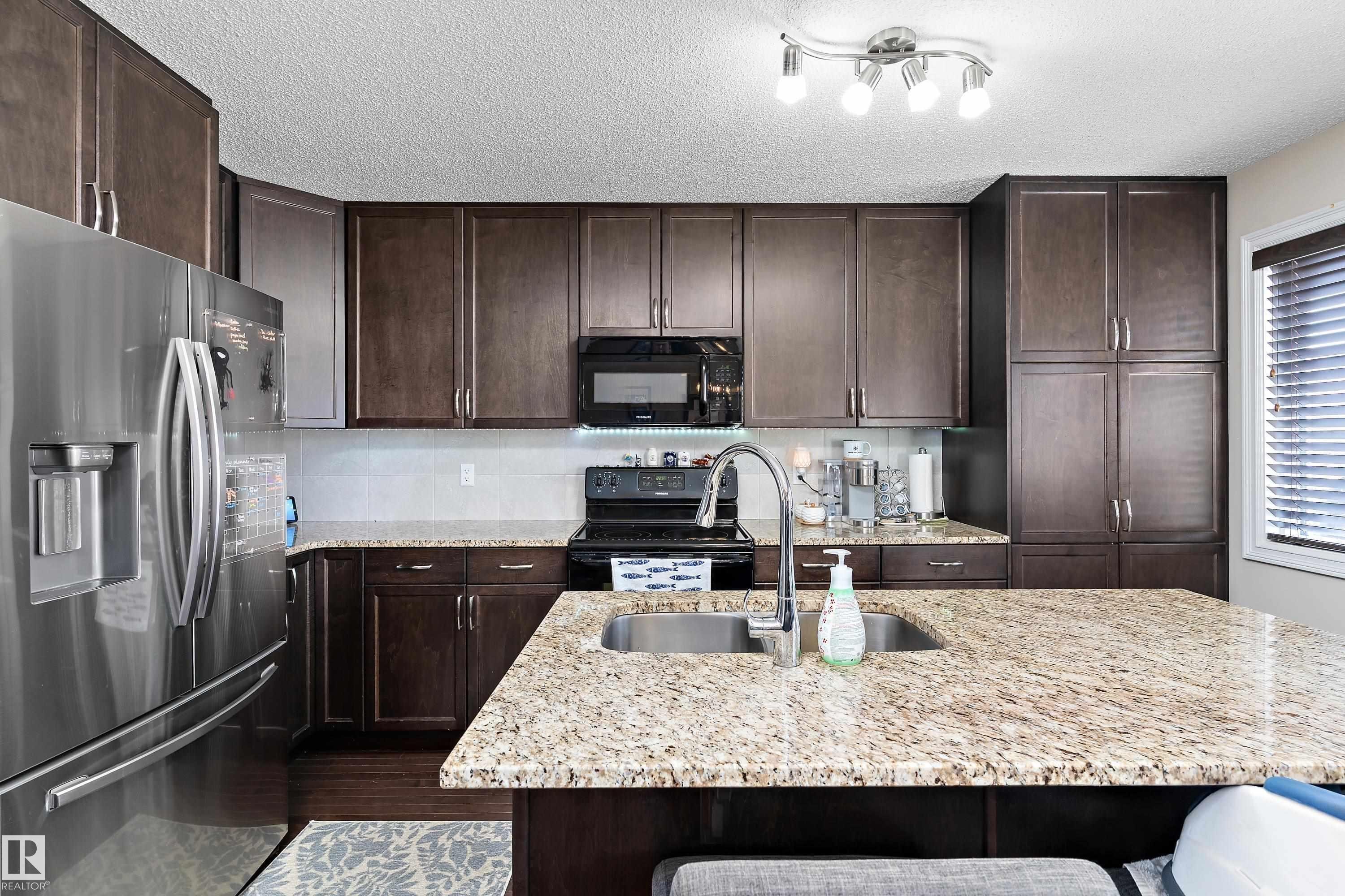 This kitchen features dark wood cabinetry, stainless steel appliances, and a granite-topped island with an integrated sink - 6822 Cardinal Link, Edmonton, AB - Indoor Photo Showing Kitchen With Upgraded Kitchen