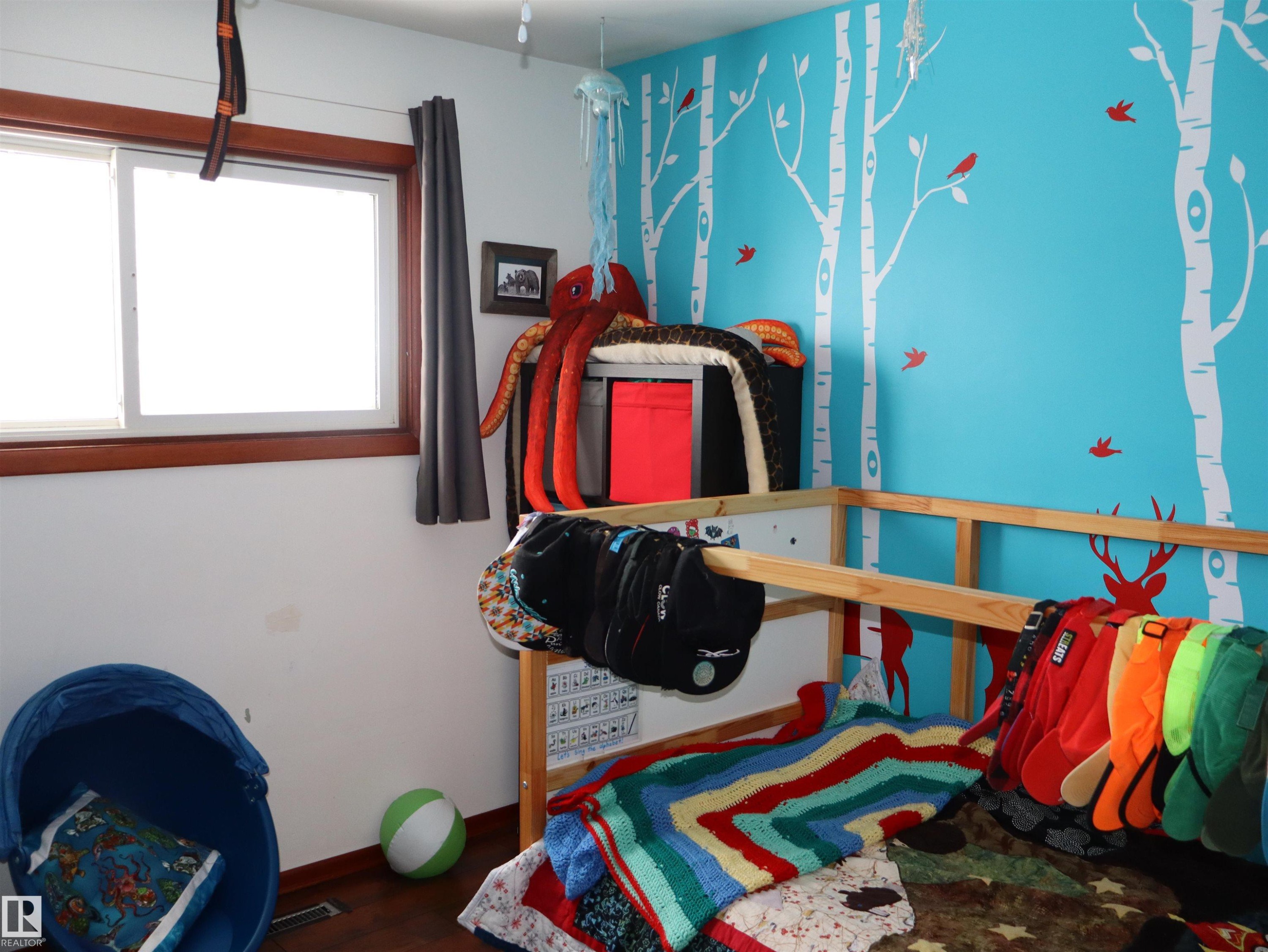 This room features a window with wood trim and dark curtains, a vibrant blue accent wall with white tree and red bird decals, and hardwood flooring - 4315 56 Avenue, Barrhead, AB - Indoor Photo Showing Bedroom