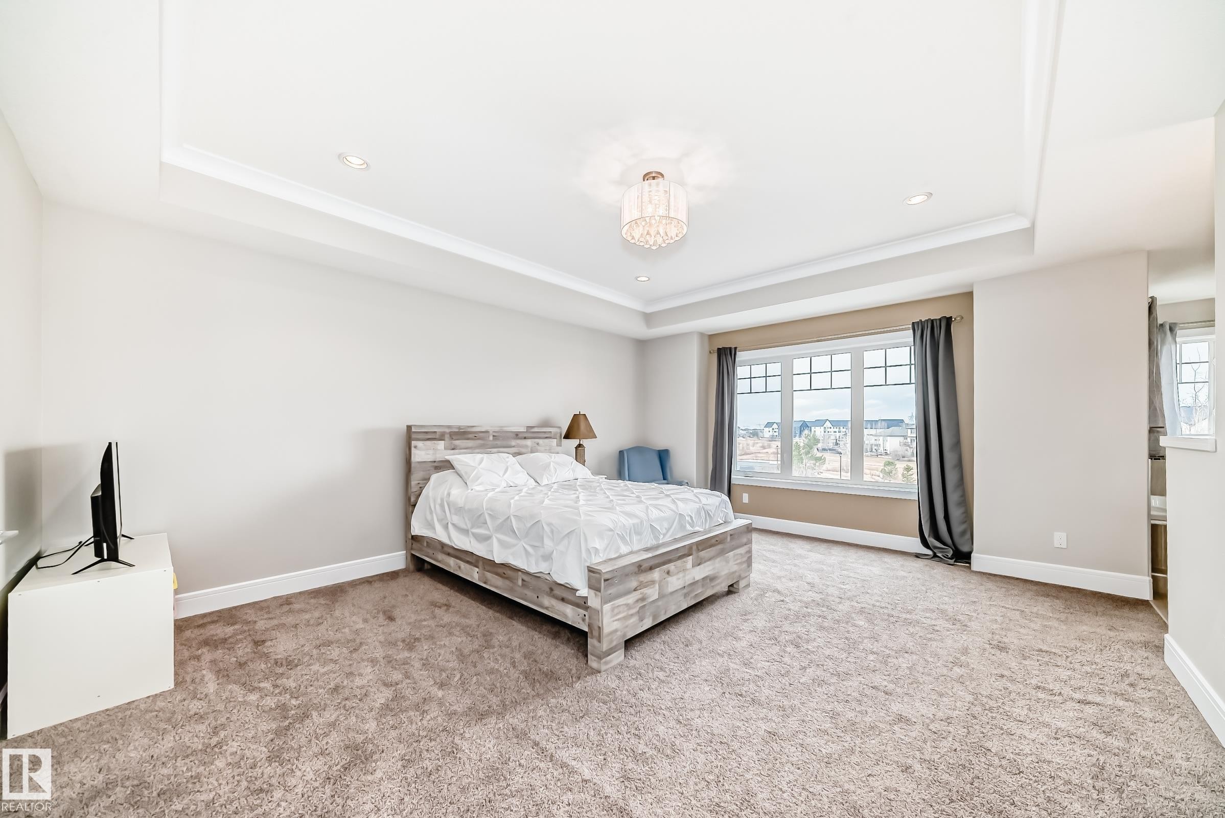 The room features a tray ceiling with recessed lighting and a chandelier, carpeted flooring, and a large window with multiple panes - 4134 Charles Link, Edmonton, AB - Indoor Photo Showing Bedroom