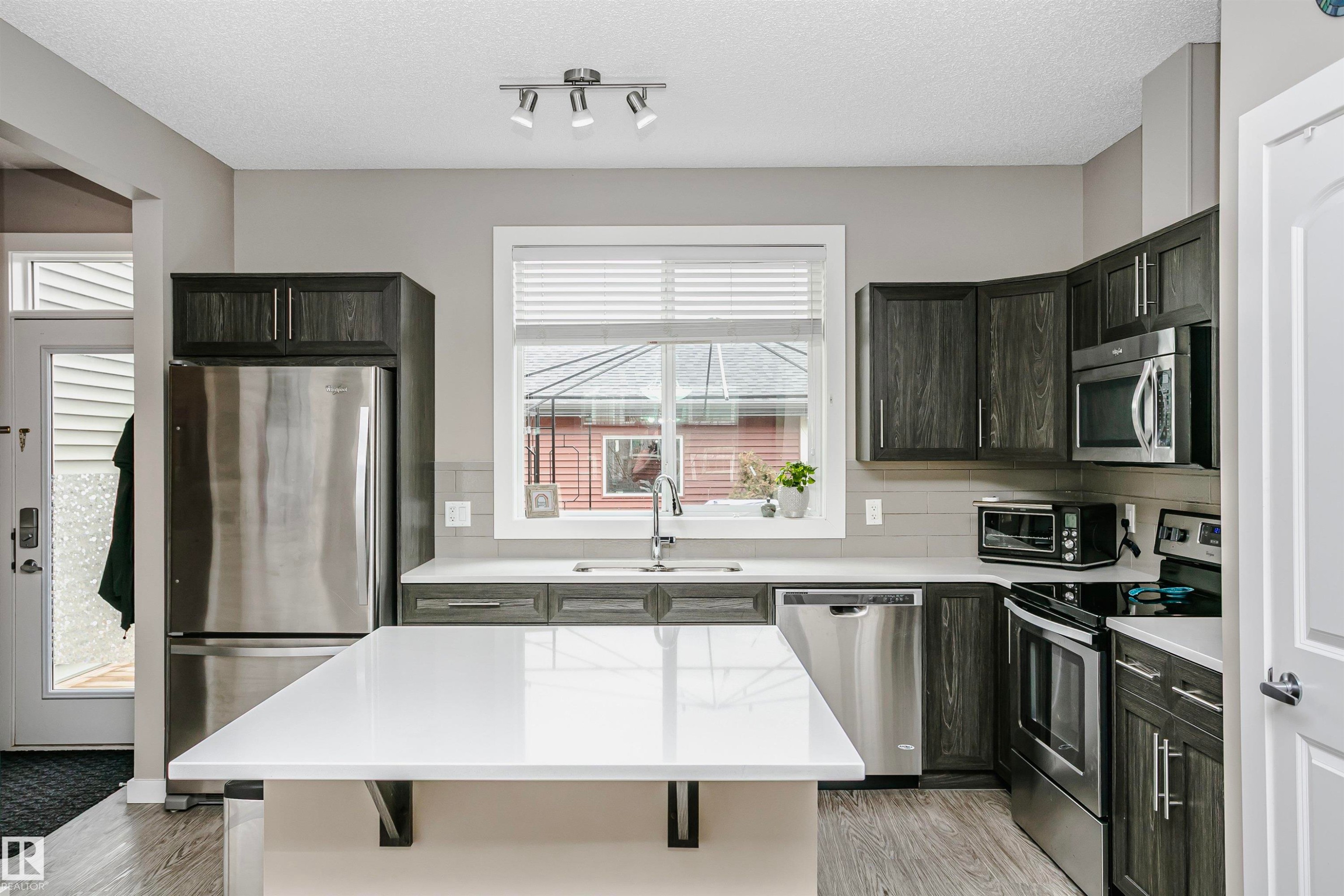 The kitchen features dark wood cabinetry, stainless steel appliances, a white island with a countertop, and a window above the sink - 6116 Rosenthal Way, Edmonton, AB - Indoor Photo Showing Kitchen With Stainless Steel Kitchen With Upgraded Kitchen
