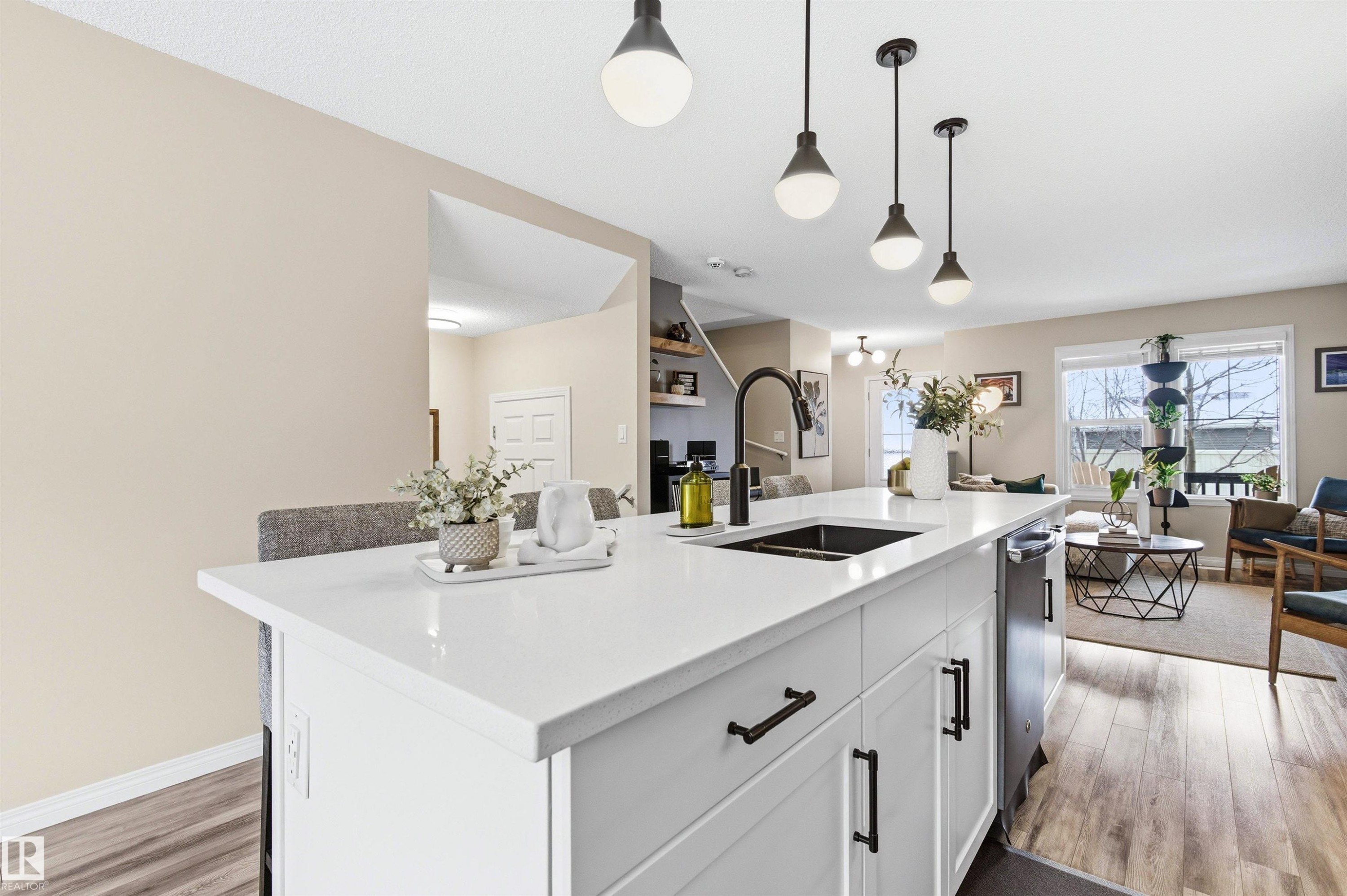 The kitchen features a white island with a sink and modern pendant lighting, overlooking a living area with hardwood style flooring and a large window - 3247 Cherry Crescent, Edmonton, AB - Indoor Photo Showing Kitchen With Double Sink
