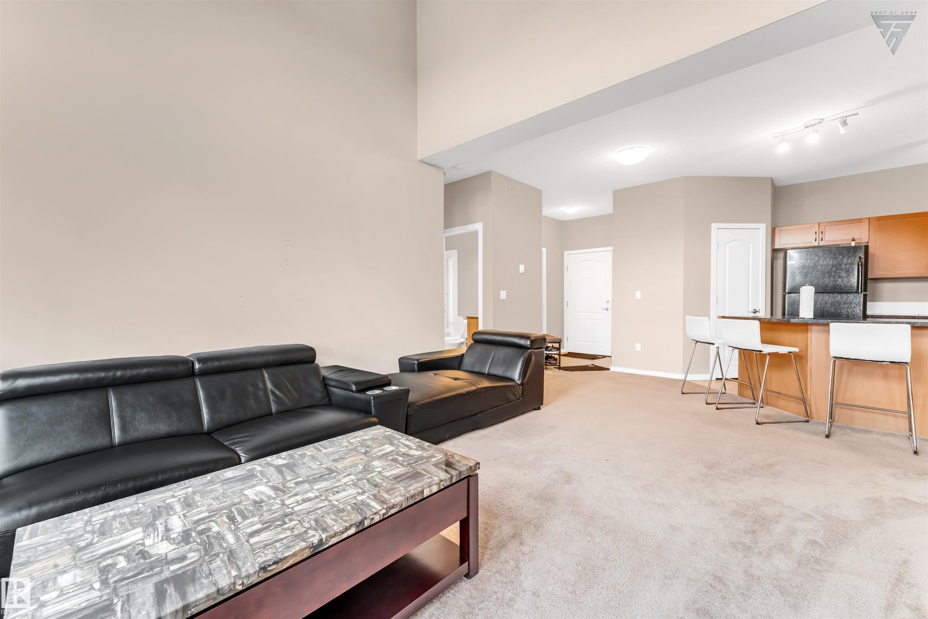 The open concept interior features light-colored walls, carpeted flooring, and a kitchen area with wood cabinetry and a black refrigerator - #418 392 Silver Berry Road Nw, Edmonton, AB - Indoor