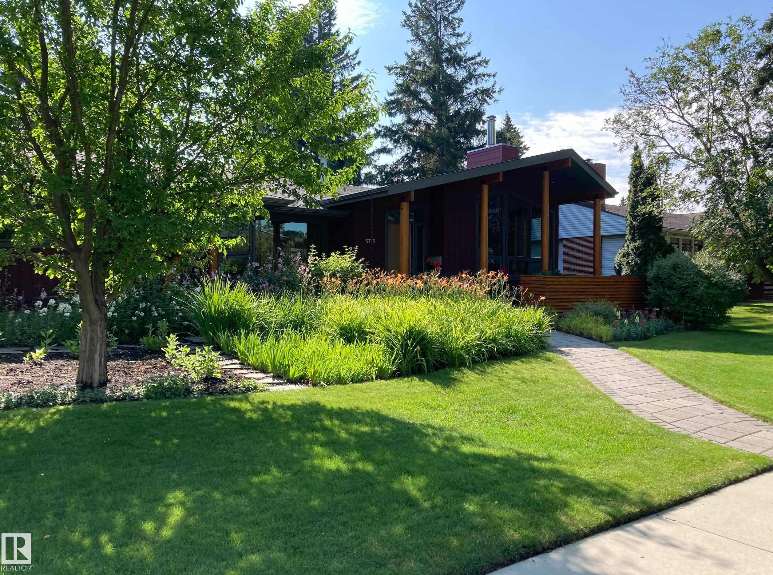 The property features a well-maintained lawn, mature trees, and a landscaped front garden with various plants and flowers - 9715 141 Street Nw, Edmonton, AB - Outdoor With Deck Patio Veranda
