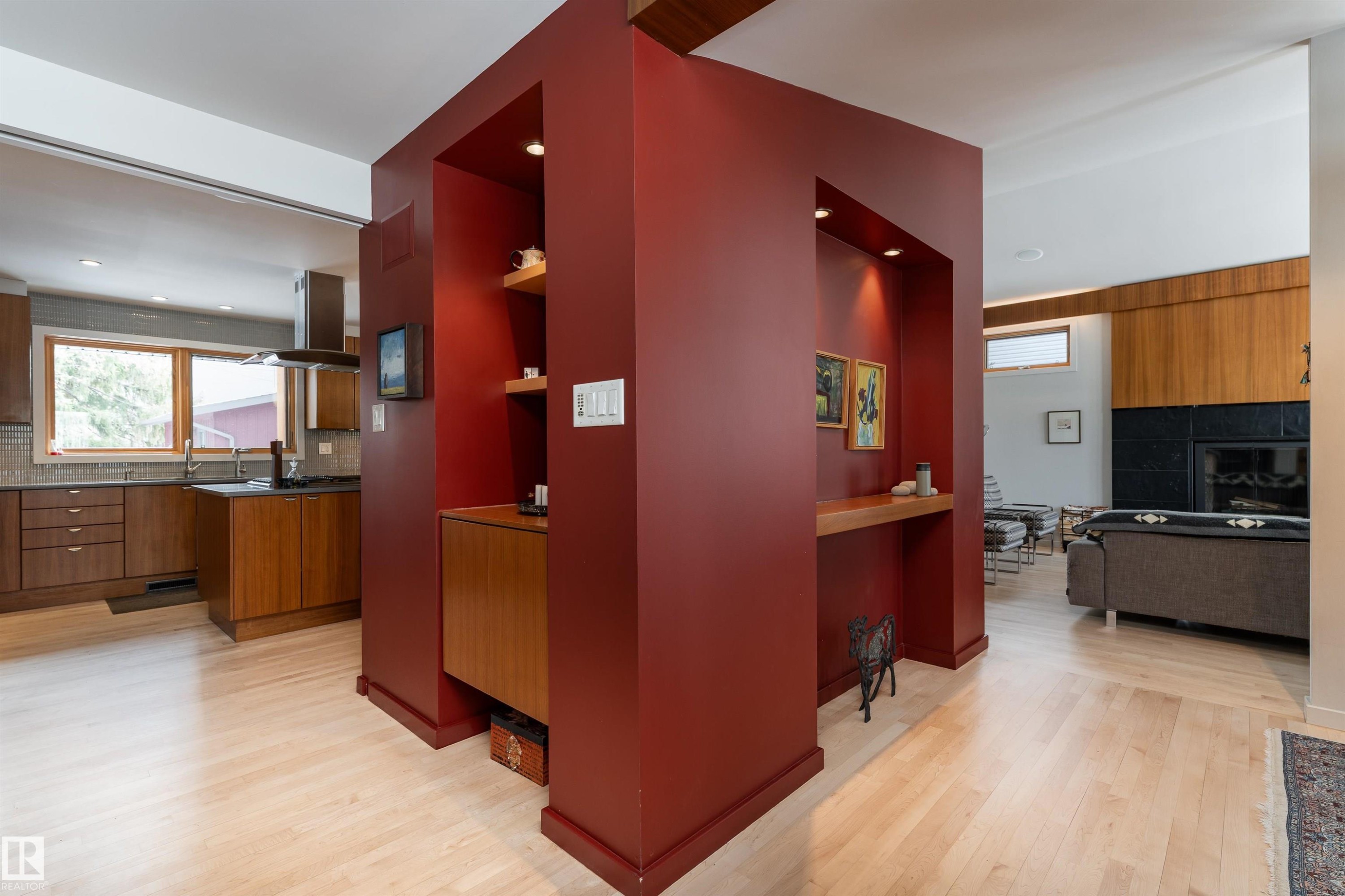 Well-lit interior featuring light-toned flooring, a red accent wall with recessed shelving and lighting, and a kitchen area with wood cabinetry and a window - 9715 141 Street Nw, Edmonton, AB - Indoor