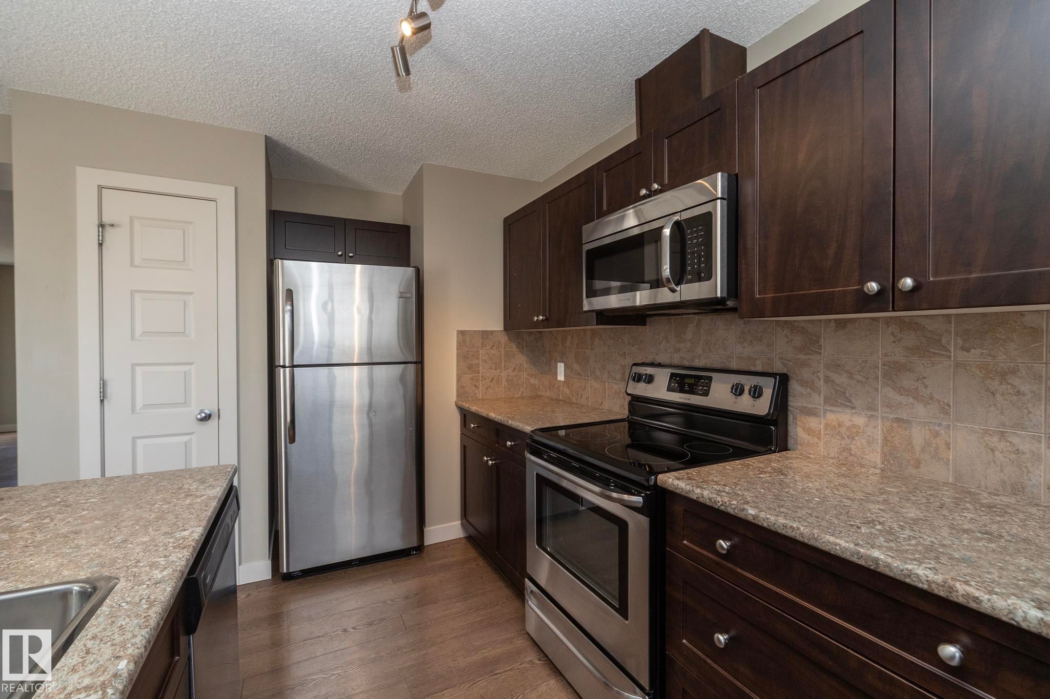 The kitchen features dark wood cabinetry, stainless steel appliances including a refrigerator, range, and microwave, and light-colored countertops with a tiled backsplash - 1403 Cunningham Drive, Edmonton, AB - Indoor Photo Showing Kitchen