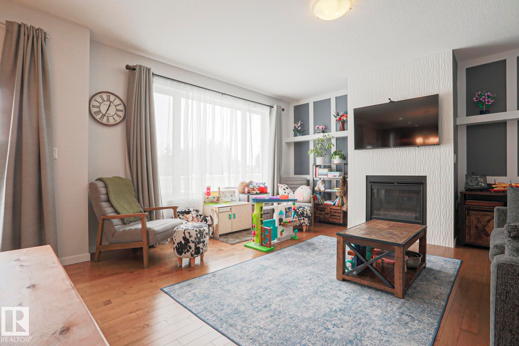 This living space features hardwood floors, a large window providing natural light, and a fireplace with a textured surround - 18 Enchanted Way, St. Albert, AB