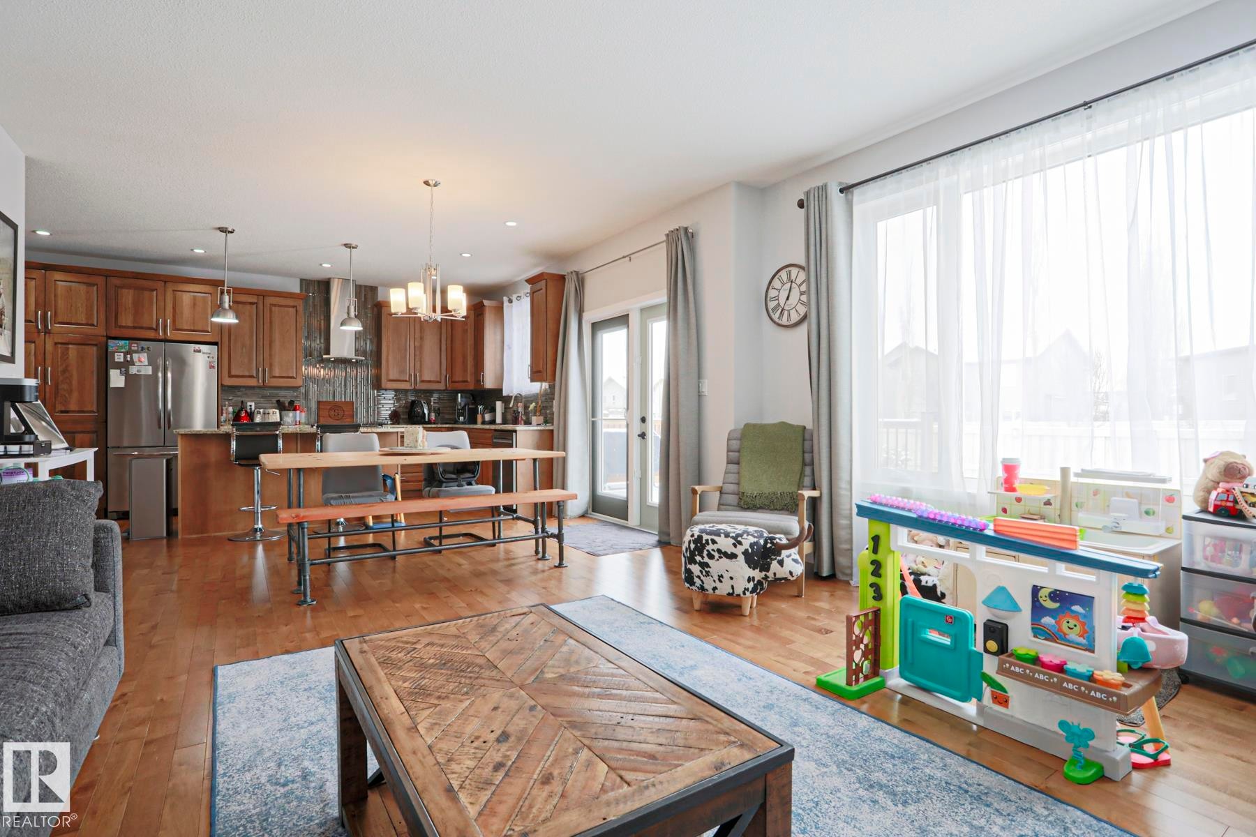 This open-concept living area features hardwood floors, a kitchen with wooden cabinetry and stainless steel appliances, and large windows providing natural light - 18 Enchanted Way, St. Albert, AB