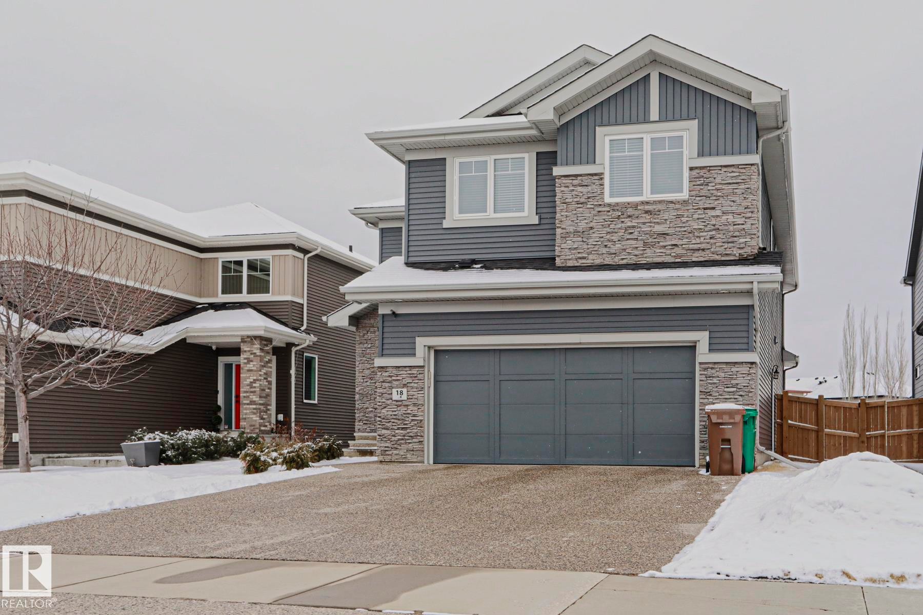 This property features a two-story exterior with grey siding and stone accents, complemented by a front-facing attached garage - 18 Enchanted Way, St. Albert, AB