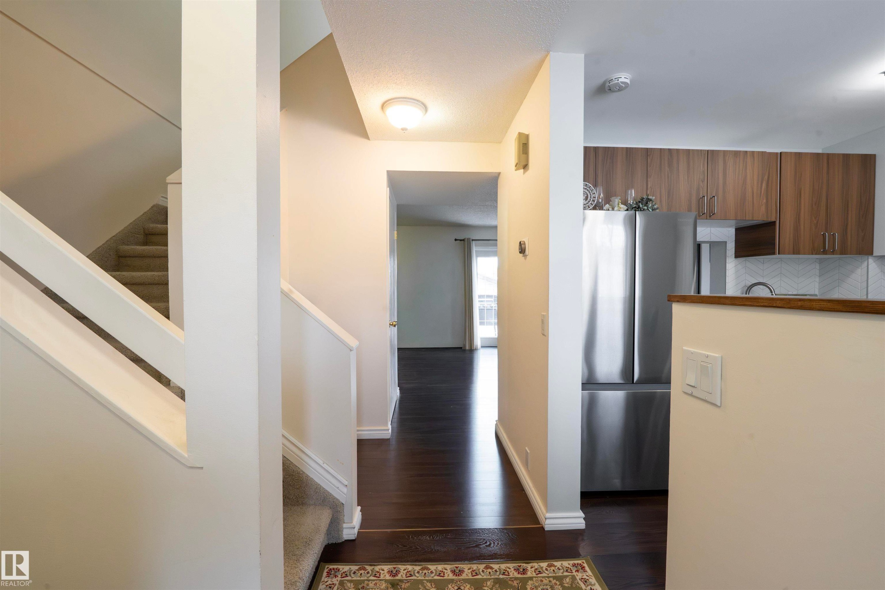 The property features dark wood flooring, a carpeted staircase with white railings, and a kitchen area with wood cabinets, a stainless steel refrigerator, and a white tile backsplash - 1728 37 Street, Edmonton, AB - Indoor