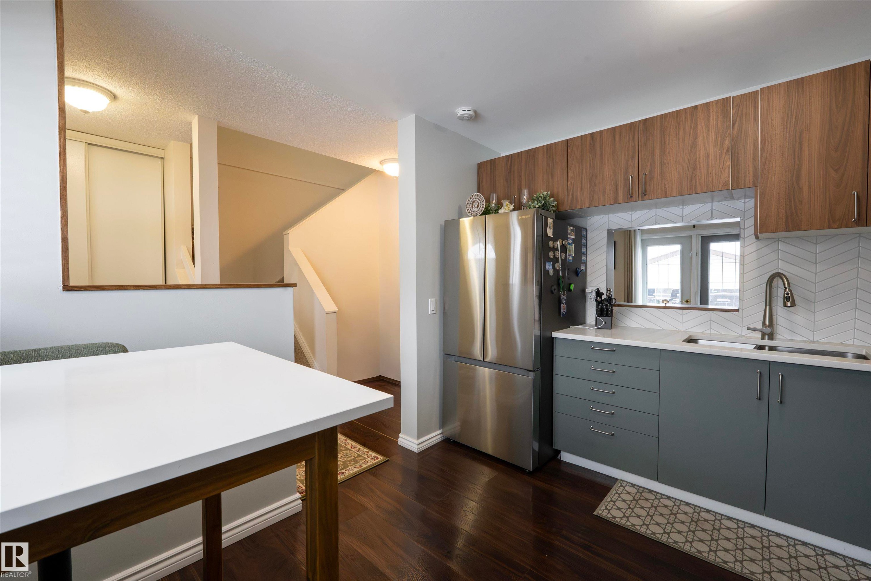 The kitchen features wood-look upper cabinetry, grey lower cabinetry, a stainless steel refrigerator, and a white quartz countertop - 1728 37 Street, Edmonton, AB - Indoor Photo Showing Kitchen With Double Sink
