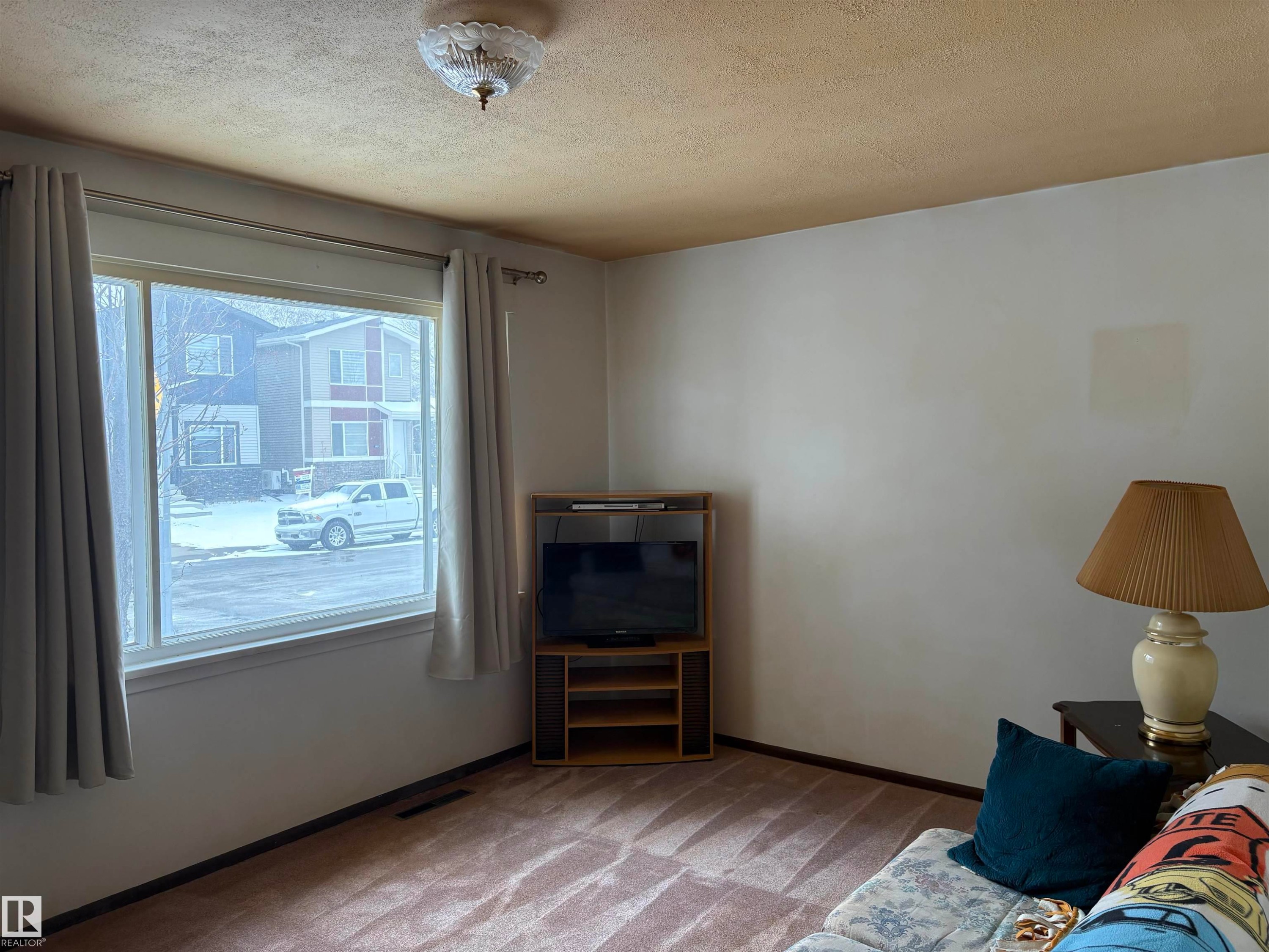 Spacious living area featuring a large window, a ceiling-mounted light fixture, and carpeted floors - 10916 151 Street, Edmonton, AB - Indoor