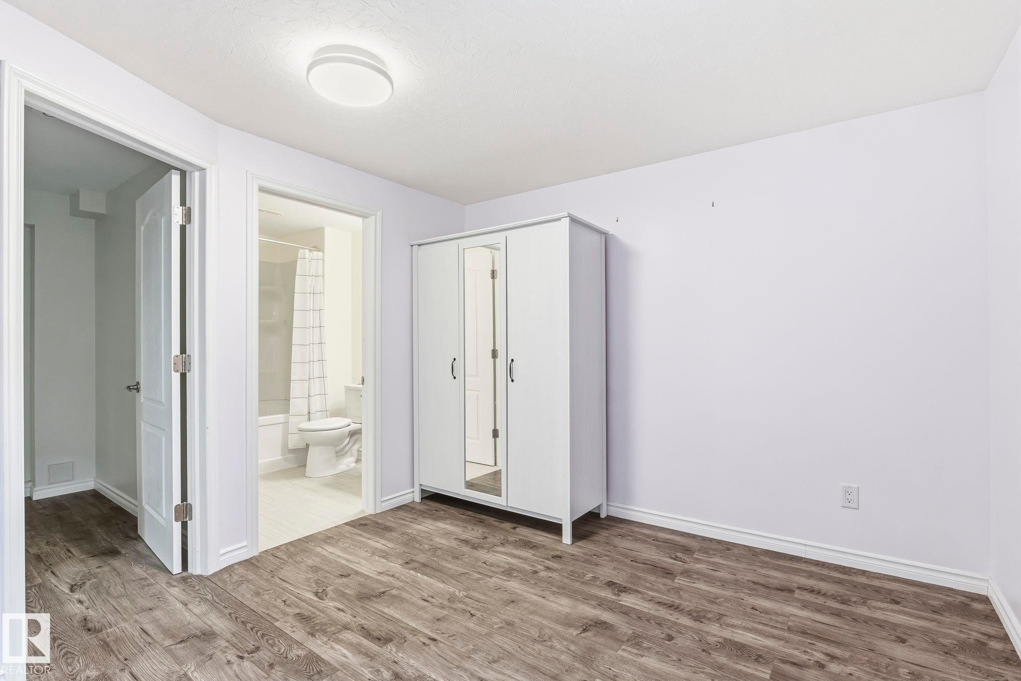 Room featuring wood-style flooring, light purple walls, a ceiling light fixture, and an open doorway leading to a bathroom with a white toilet and a shower with a patterned curtain - 4783 Crabapplle Run, Edmonton, AB - Indoor Photo Showing Other Room