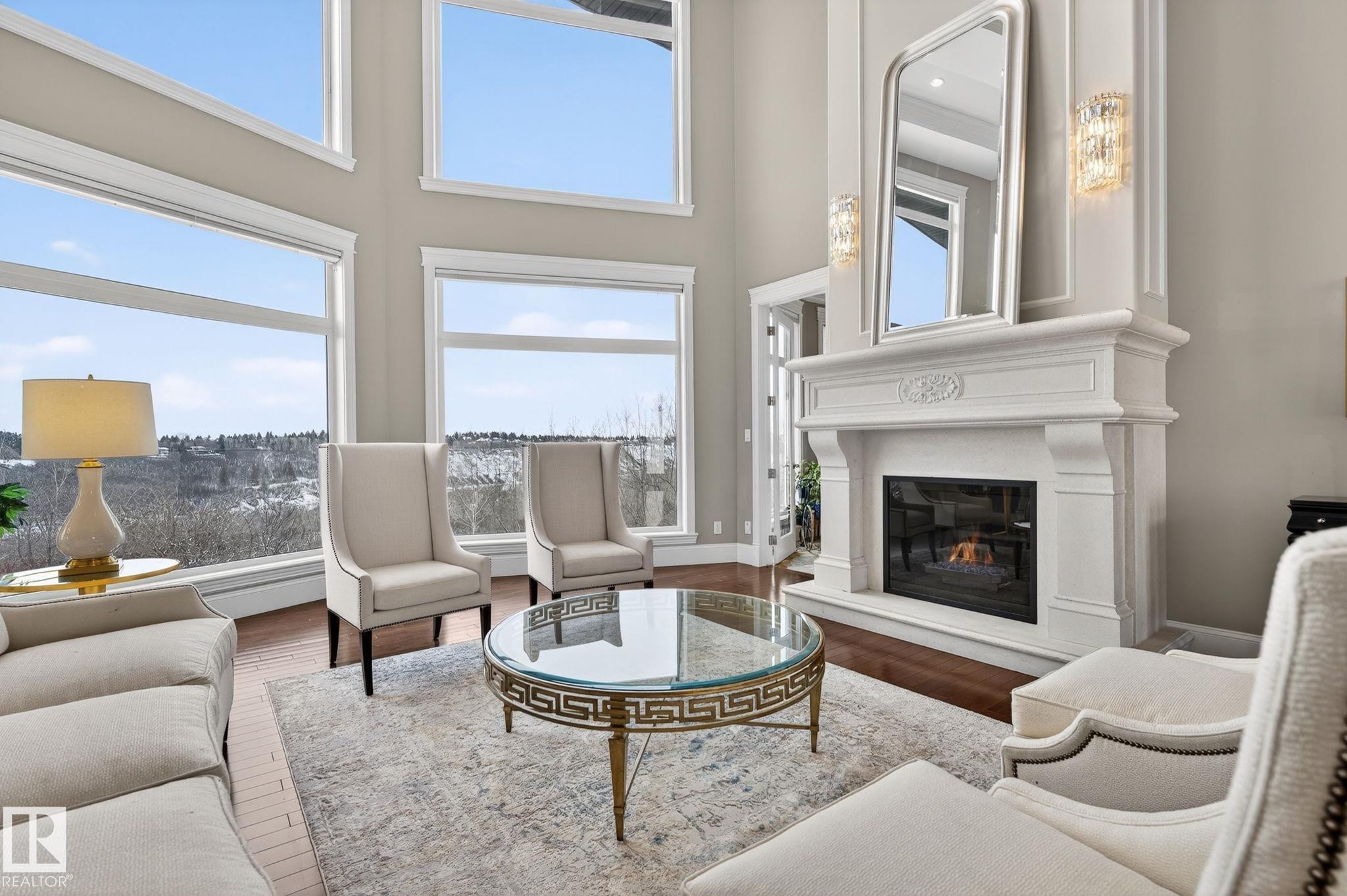 This inviting living space features high ceilings, expansive windows offering abundant natural light and exterior views, a detailed white mantel with a fireplace, and hardwood flooring - 1413 Woodward Crescent, Edmonton, AB - Indoor Photo Showing Living Room With Fireplace