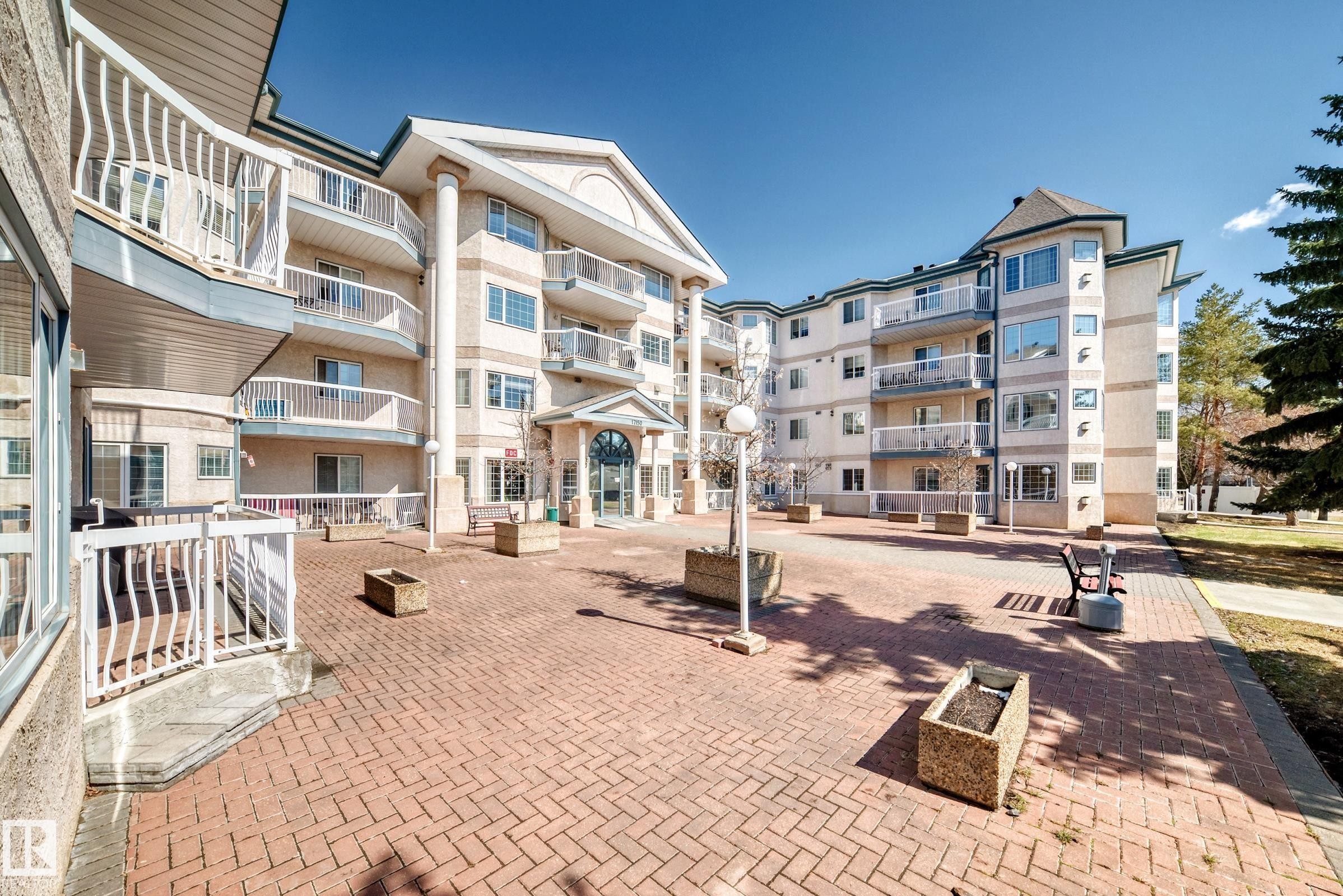 The property features a multi-story building with a light-colored exterior and multiple balconies, set on a brick paved courtyard - 112 17150 94A Avenue, Edmonton, AB - Outdoor