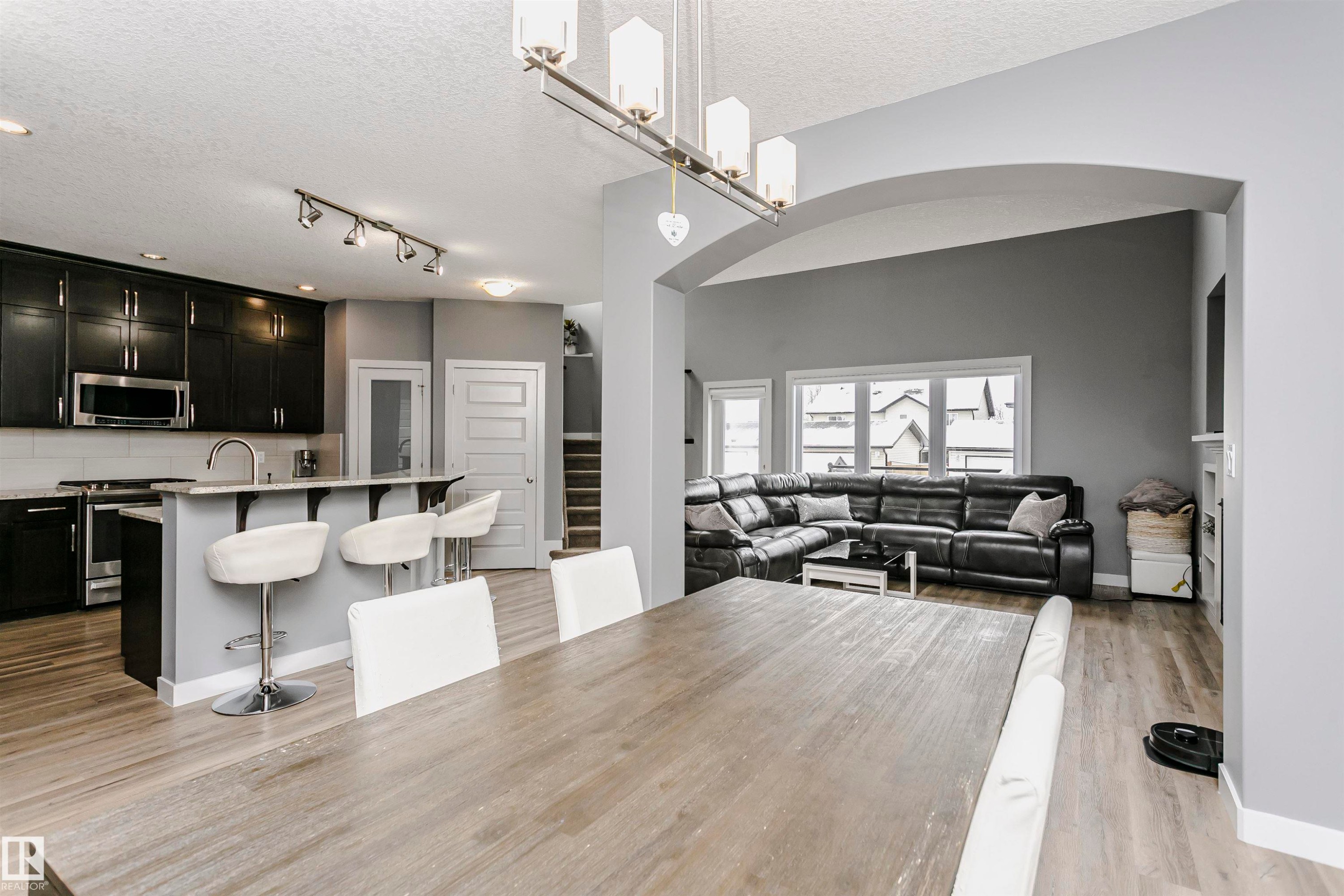 Open concept living space featuring a dining area with a wood-toned table, an archway leading to a living room with large windows, and a kitchen with dark cabinetry, a breakfast bar, and stainless steel appliances - 21807 97 Avenue, Edmonton, AB - Indoor