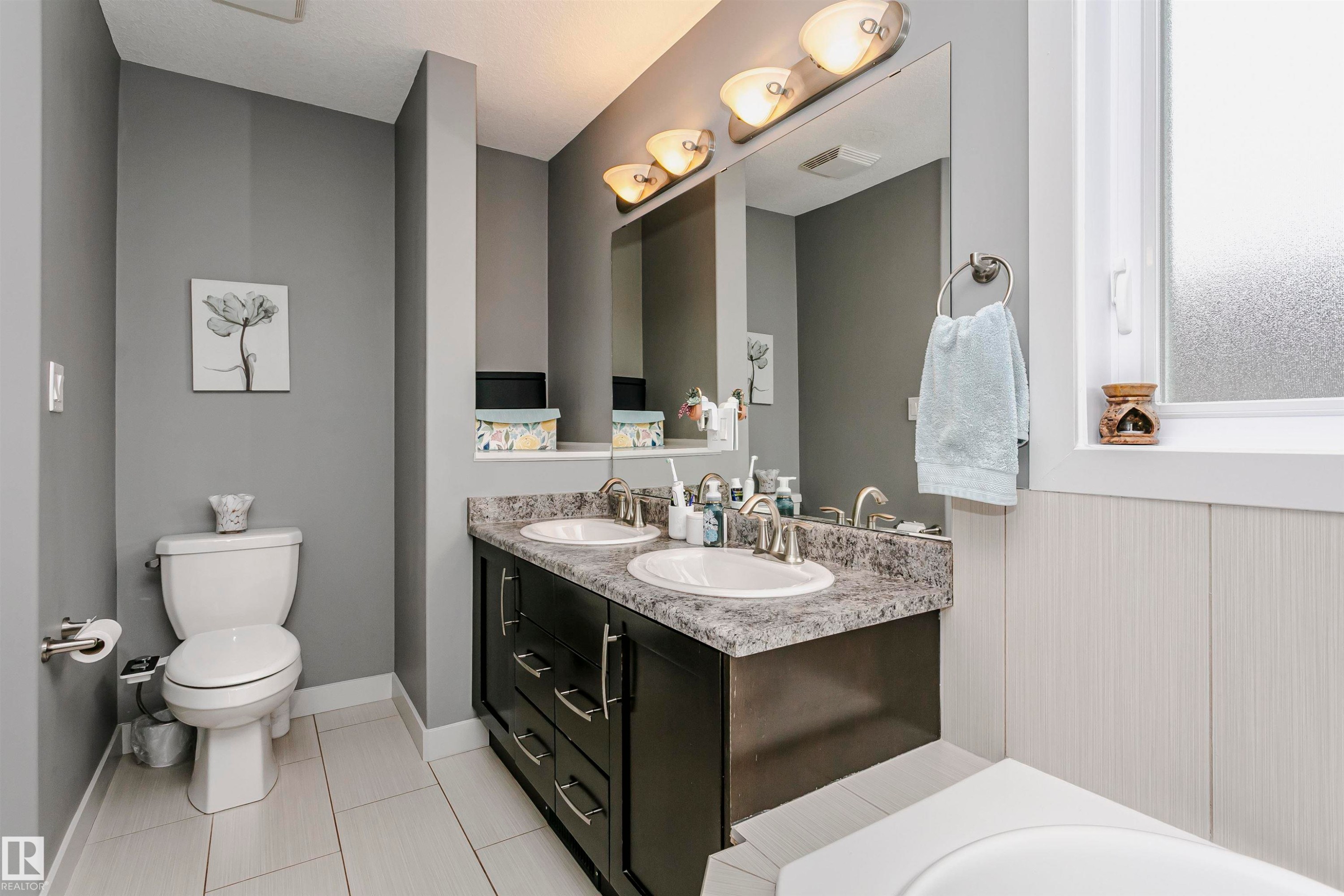 The bathroom features a dual sink vanity with dark cabinetry, a toilet, a window, and light-colored wall paneling - 21807 97 Avenue, Edmonton, AB - Indoor Photo Showing Bathroom
