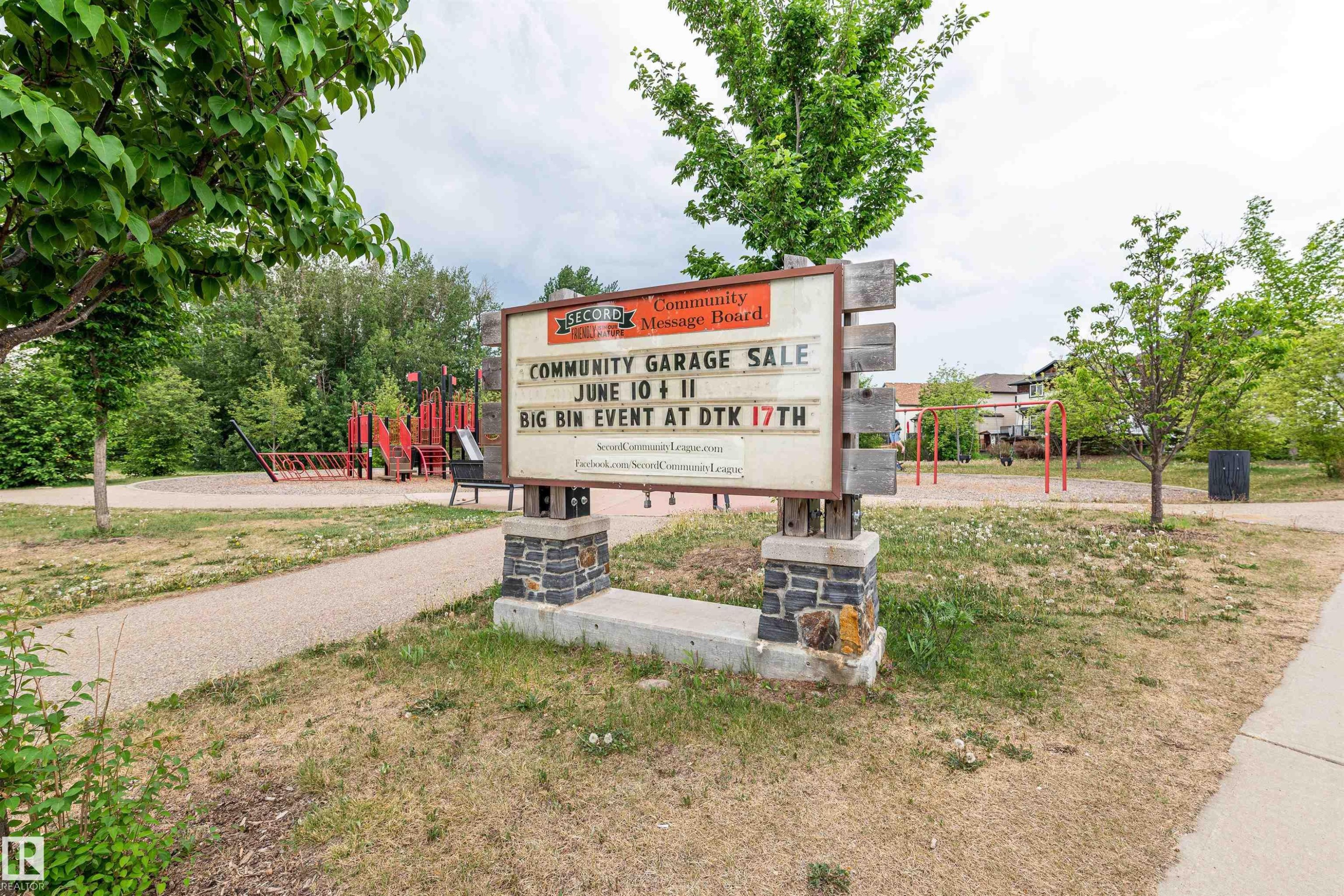 Community message board on a stone and wood base, set within a park area featuring green trees and a paved pathway - 21807 97 Avenue, Edmonton, AB - Outdoor