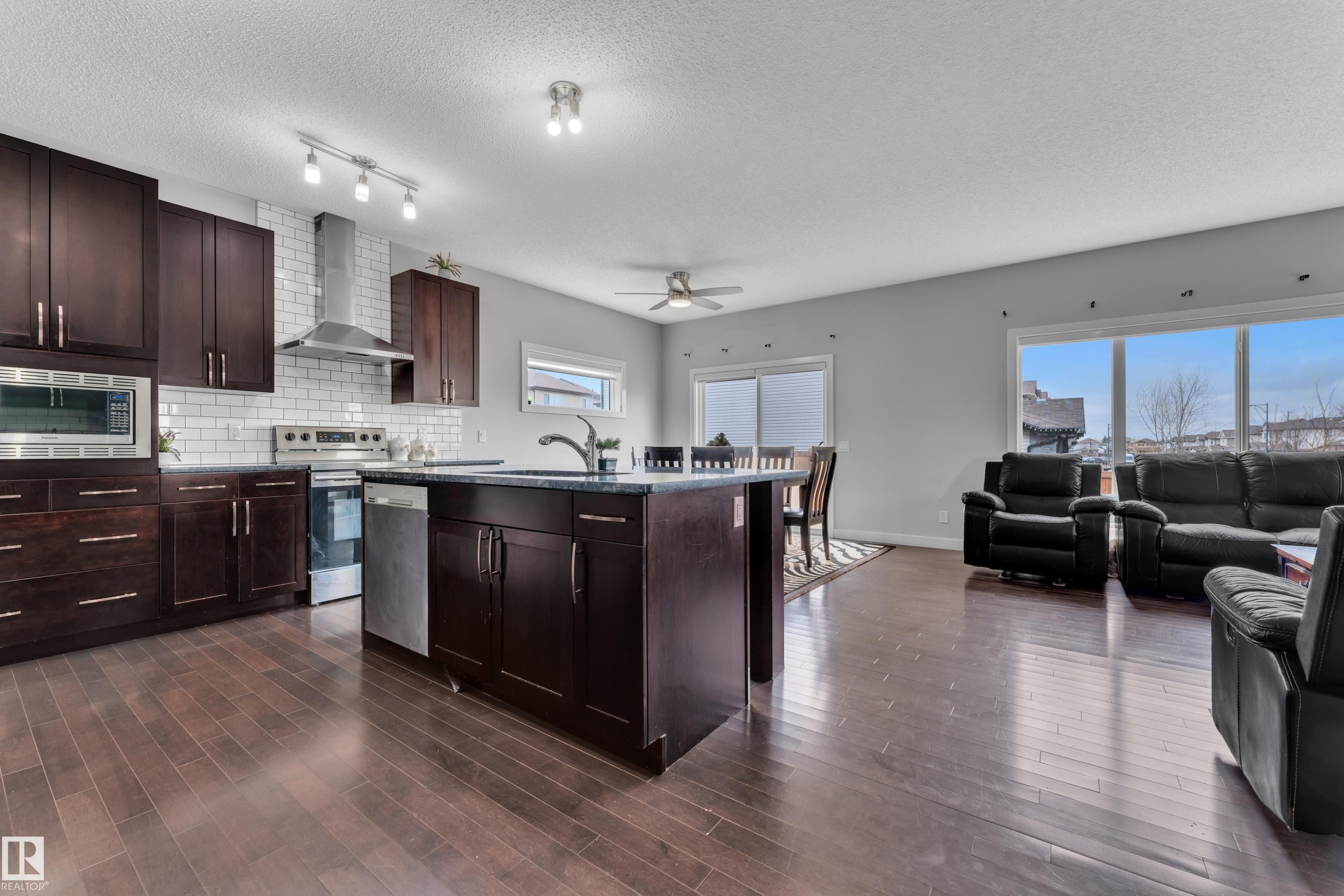 Open concept living area featuring dark hardwood flooring, a kitchen with dark cabinetry, stainless steel appliances, and a white subway tile backsplash, and large windows providing ample natural light - 1452 30 Street, Edmonton, AB - Indoor Photo Showing Kitchen With Upgraded Kitchen