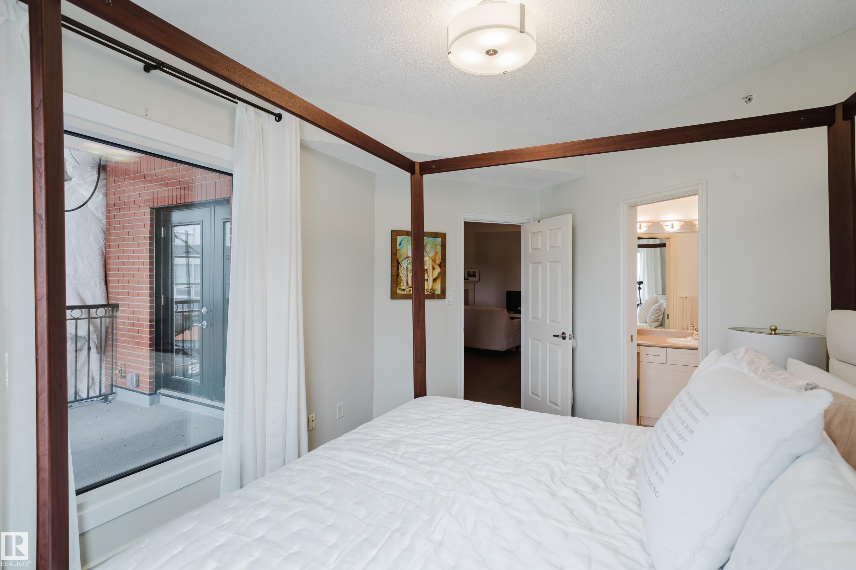 The bedroom features white walls, a ceiling-mounted light fixture, and a large glass door providing access to a balcony with a black wrought iron railing and a brick exterior wall - 206 10108 125 Street, Edmonton, AB - Indoor Photo Showing Bedroom