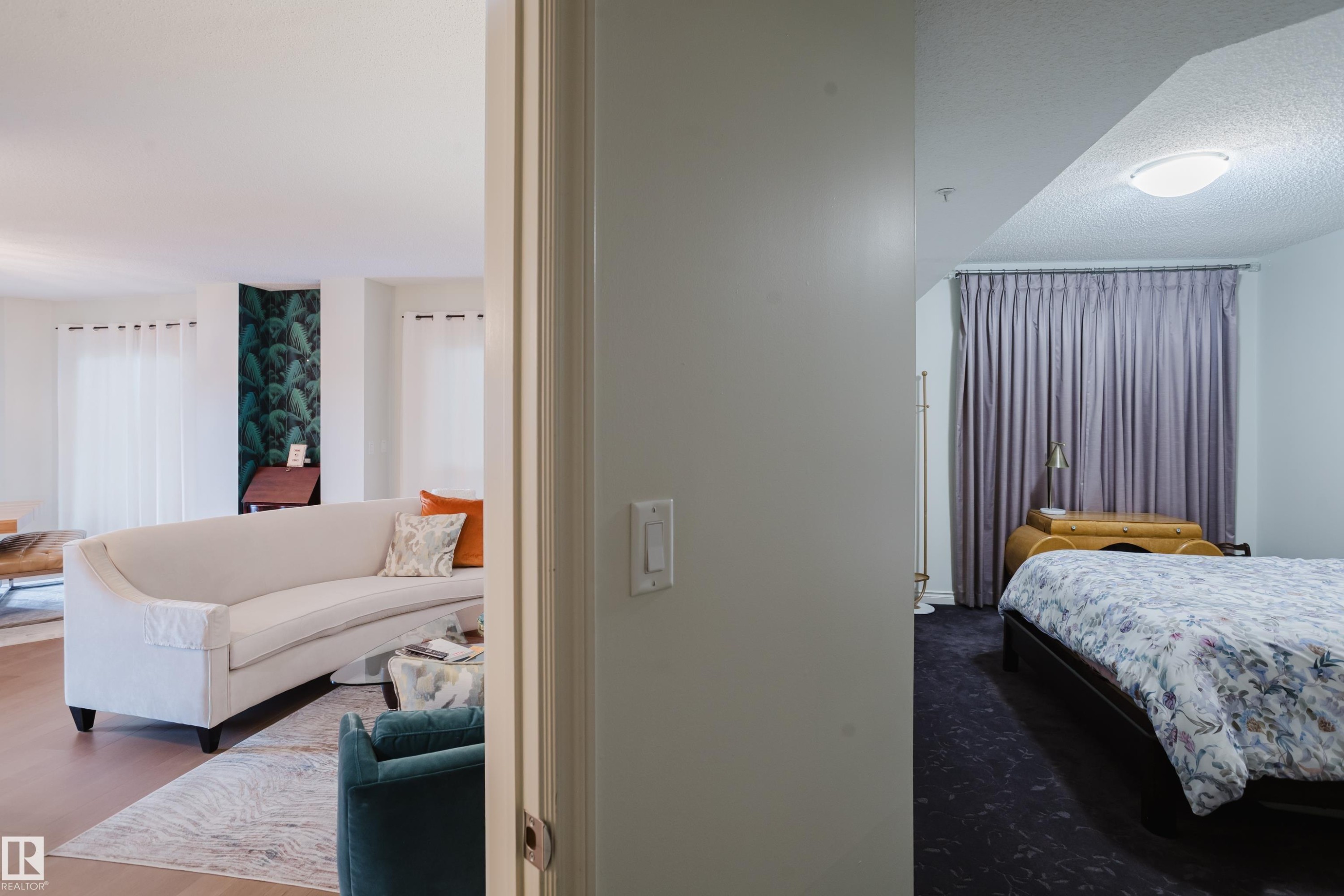 The property features a bright living area with light hardwood floors and a decorative accent wall, visible from a separate room with dark flooring and privacy curtains - 206 10108 125 Street, Edmonton, AB - Indoor Photo Showing Bedroom