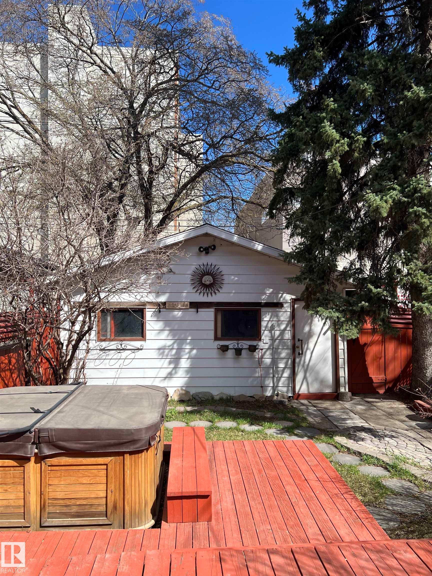 Expansive wood deck with integrated seating, white siding exterior building with dual windows, and mature evergreen trees - 10918 81 Avenue, Edmonton, AB - Outdoor