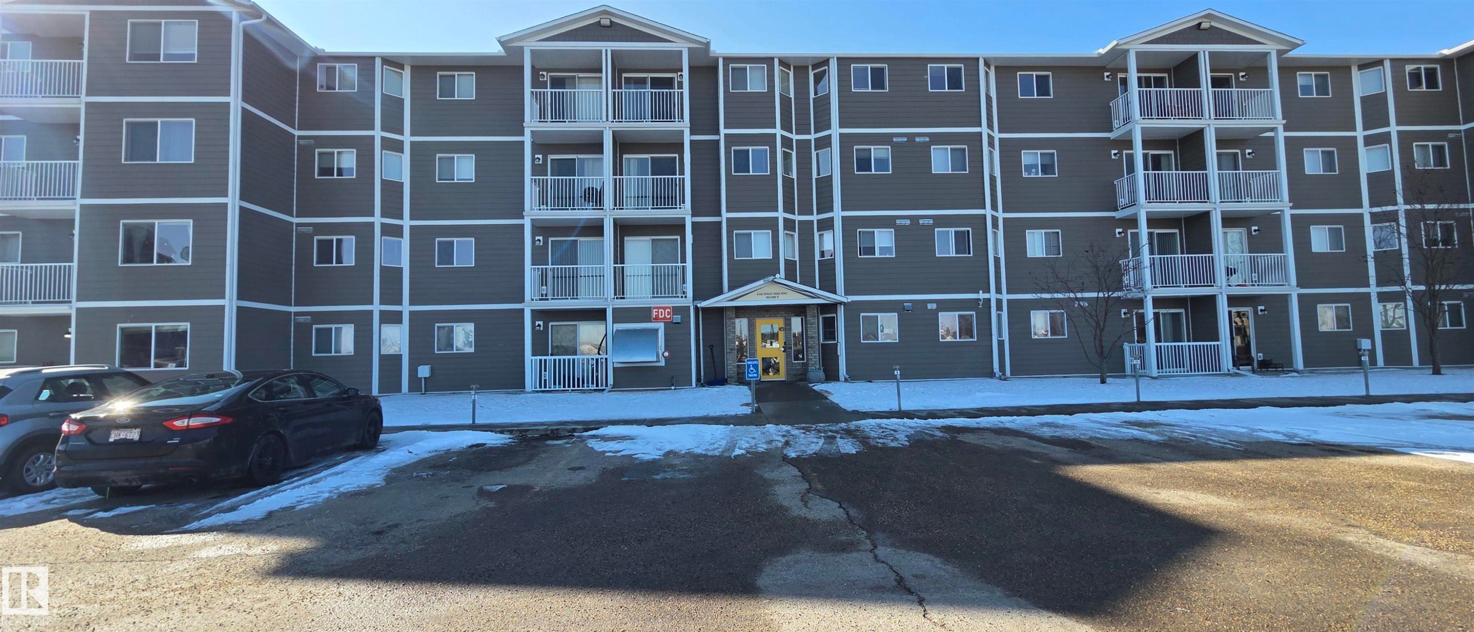 The property features a five-story building with balconies, a paved parking area, and a central entrance - 208A 260 Spruce Ridge Road, Spruce Grove, AB - Outdoor With Balcony With Facade