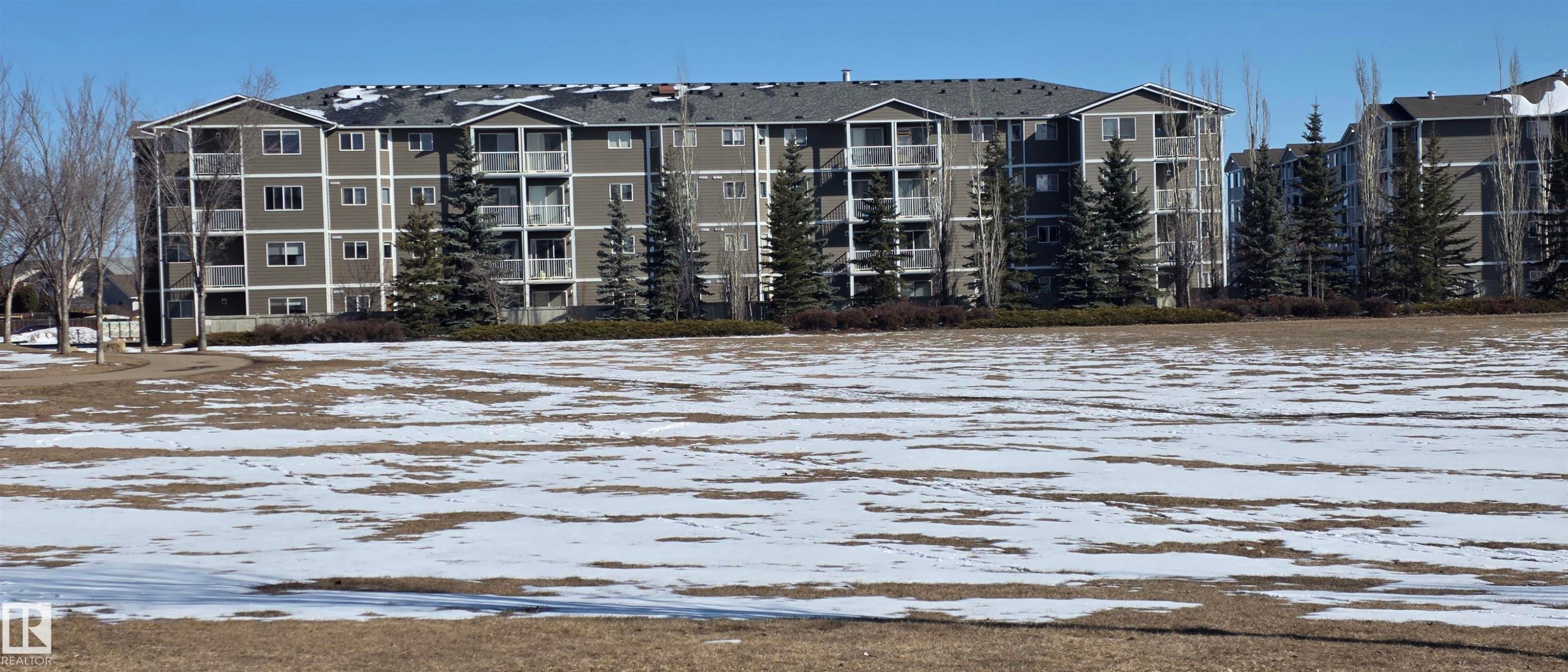 The property features a multi-story building with balconies, a dark-colored roof, and a light-colored exterior facade - 208A 260 Spruce Ridge Road, Spruce Grove, AB - Outdoor With Body Of Water