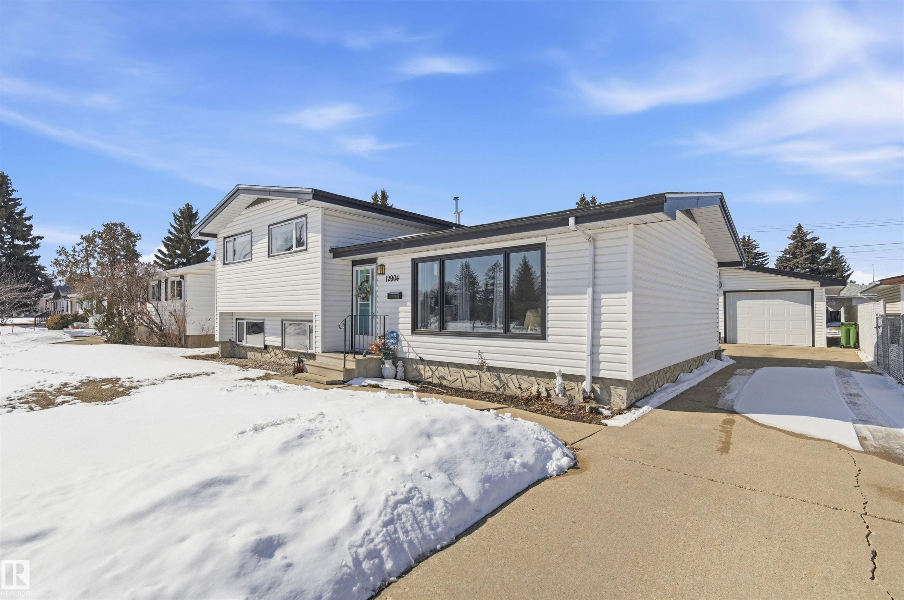 The property features a white exterior with dark trim windows, a front entrance with a light blue door, and a concrete driveway leading to a garage - 12904 95 Street, Edmonton, AB - Outdoor