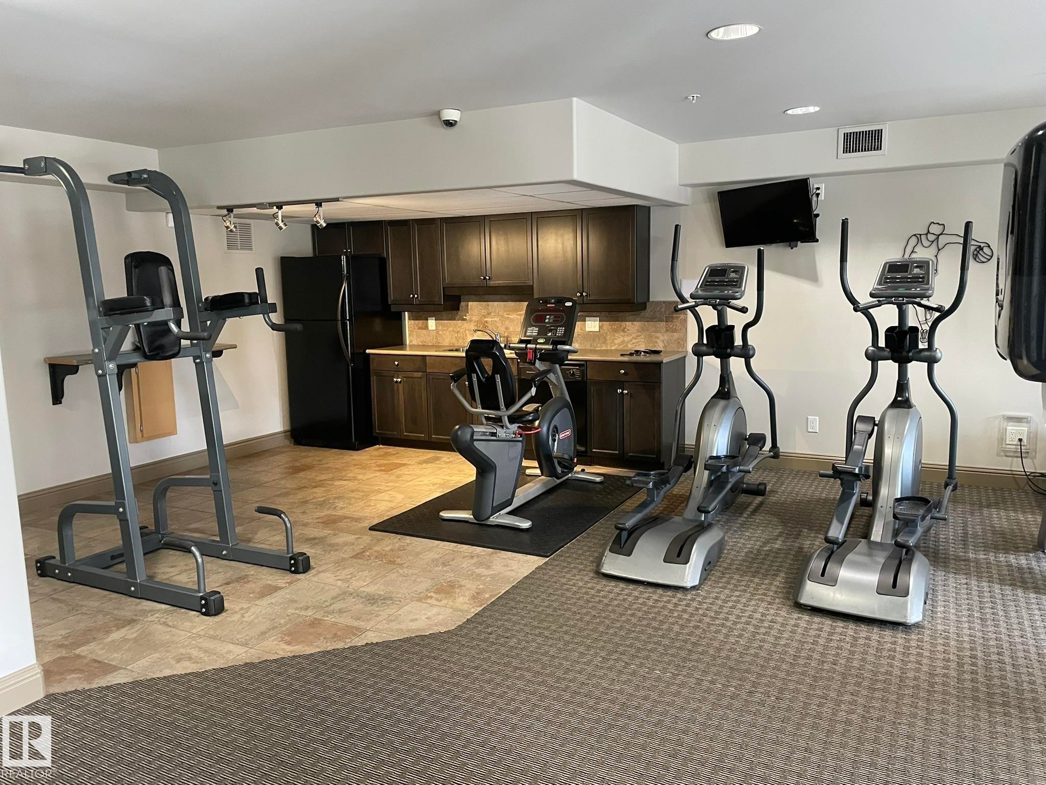 This fitness area features a variety of exercise equipment and a kitchenette with dark wood cabinetry and a black refrigerator - Edmonton, AB - Indoor Photo Showing Gym Room