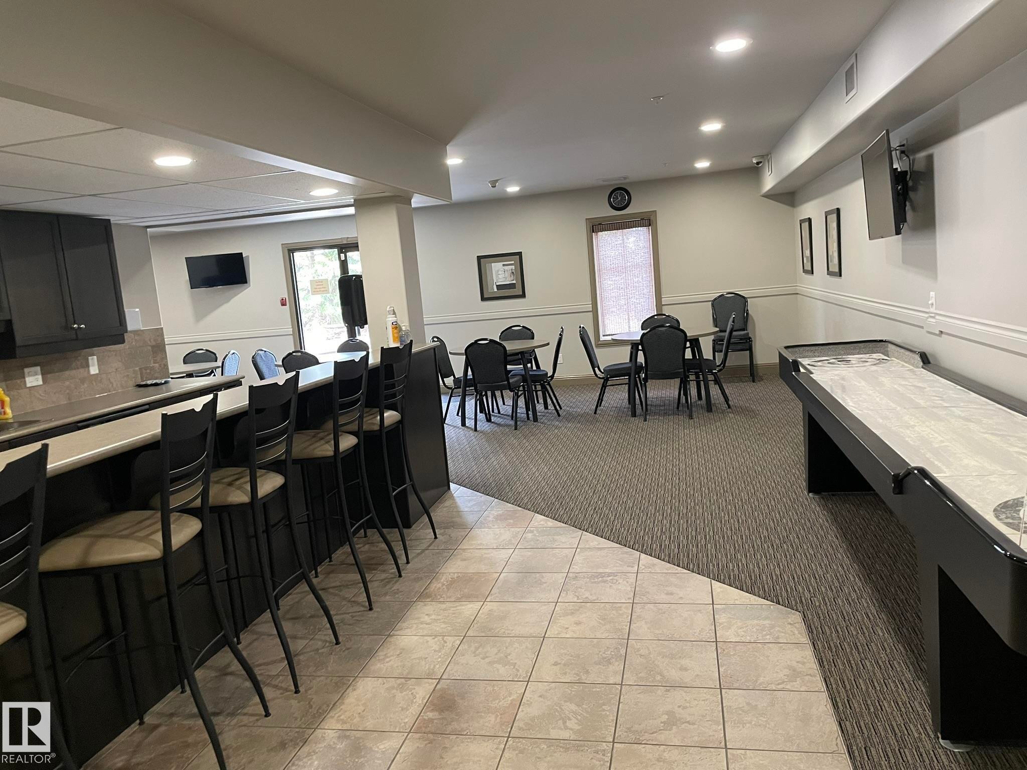 This spacious room features a tiled floor area leading to a carpeted section, a bar with seating, and a shuffleboard table - Edmonton, AB - Indoor