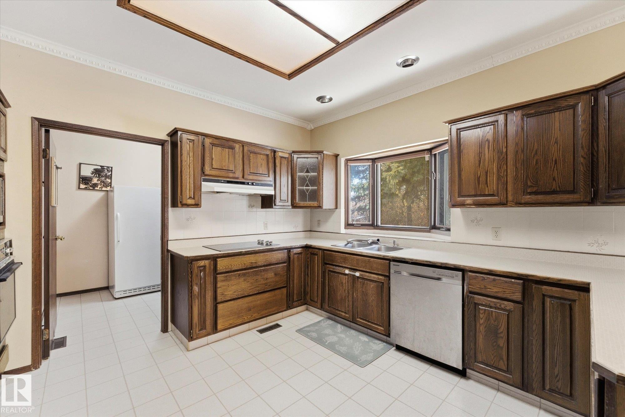 U-shaped kitchen featuring wood-finish cabinetry, white tile flooring, and a stainless steel dishwasher - 25750 18 Street, Edmonton, AB - Indoor Photo Showing Kitchen With Double Sink