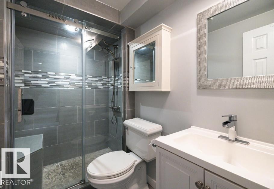 Bathroom featuring a glass-enclosed shower with tiled walls and a built-in bench, a white vanity with a rectangular sink, a toilet, and a wall-mounted mirror with a textured frame Basement bathroom - 71 Labelle Crescent, St. Albert, AB - Indoor Photo Showing Bathroom