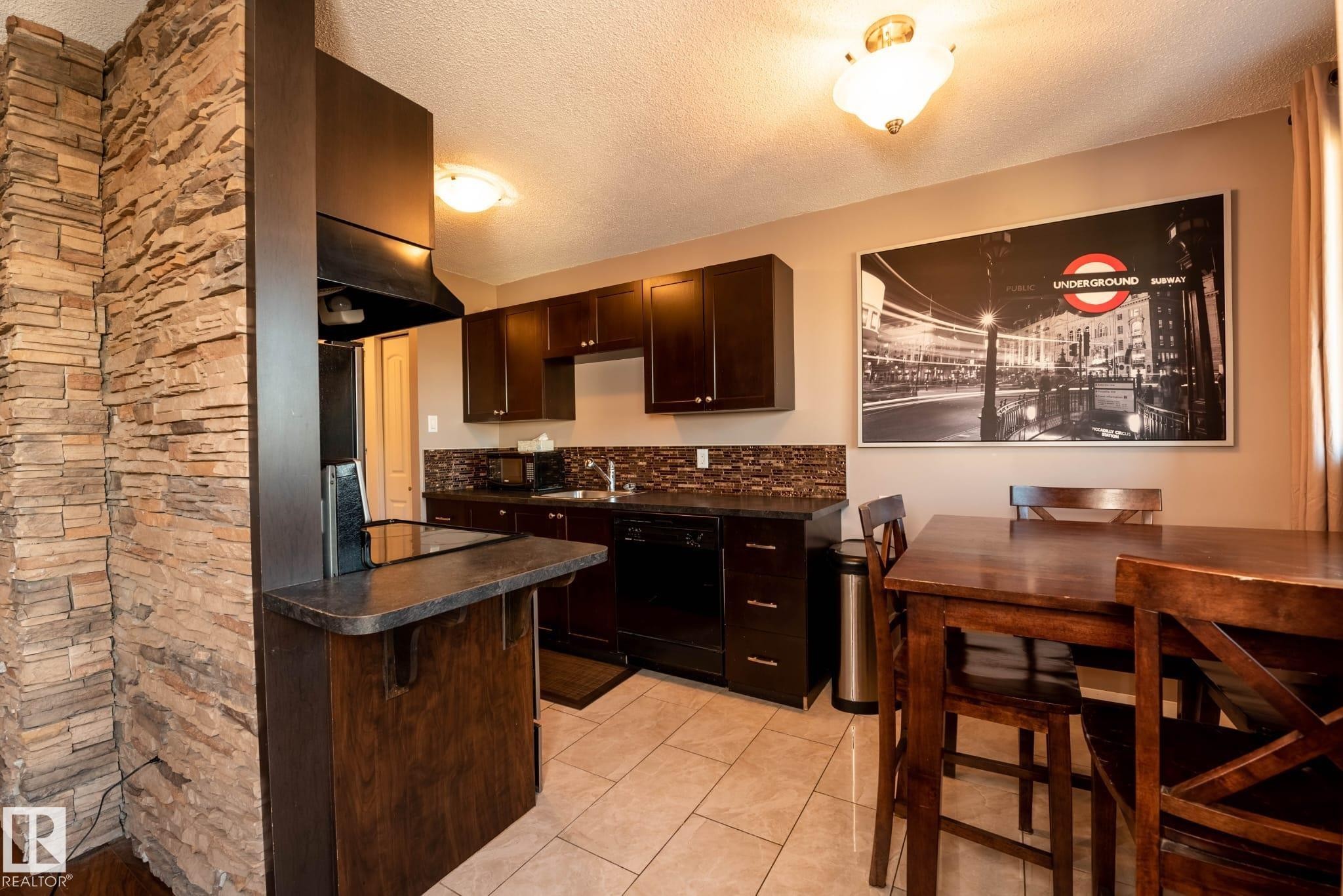 Kitchen featuring dark cabinetry, a built-in cooktop, and a stone-finish accent wall - 86 13435 97 Street, Edmonton, AB - Indoor