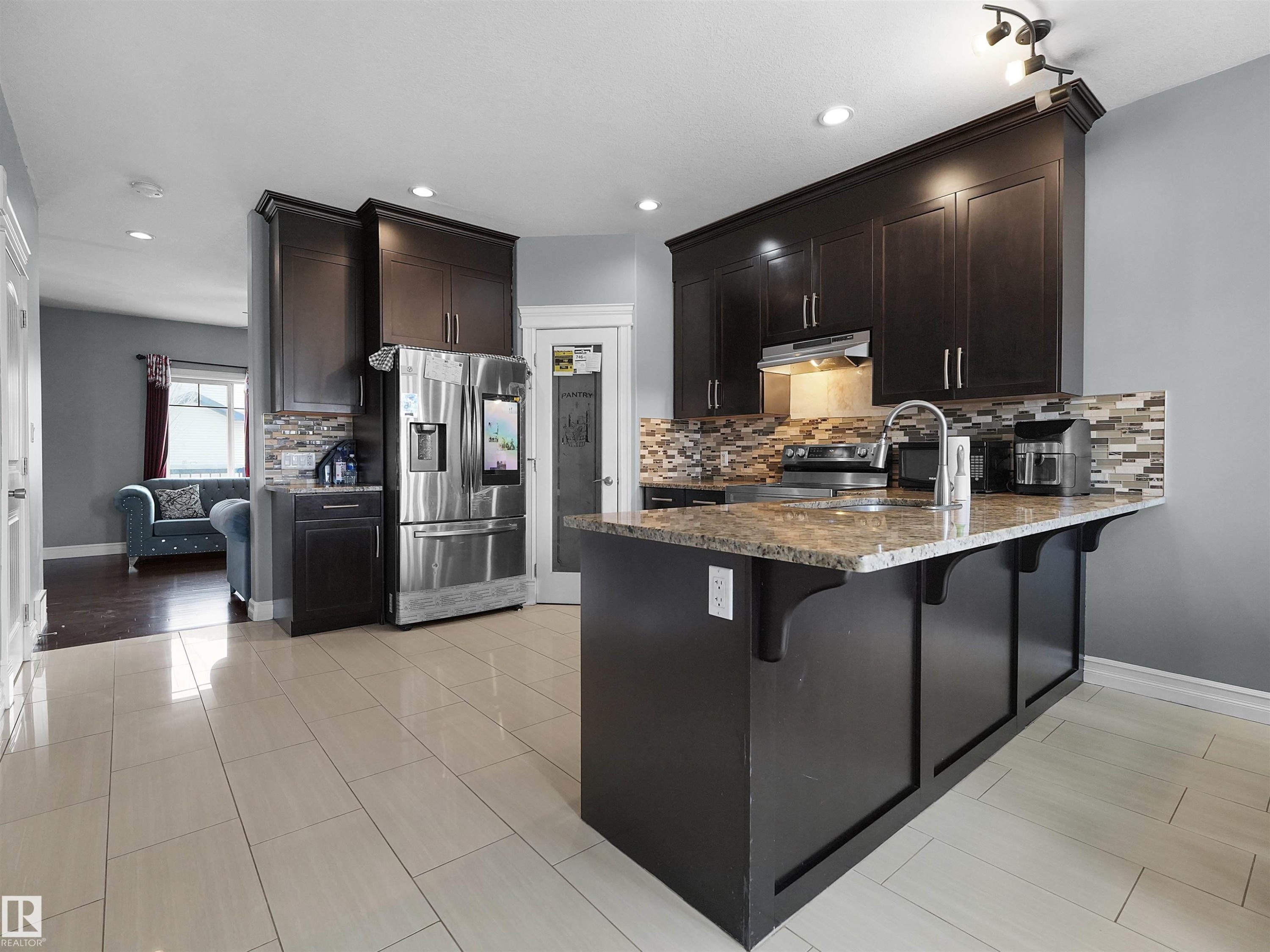 Kitchen featuring dark wood cabinetry, a granite-finish countertop island with sink, stainless steel appliances, and a mosaic tile backsplash - 5192 1B Avenue Sw, Edmonton, AB - Indoor Photo Showing Kitchen With Upgraded Kitchen