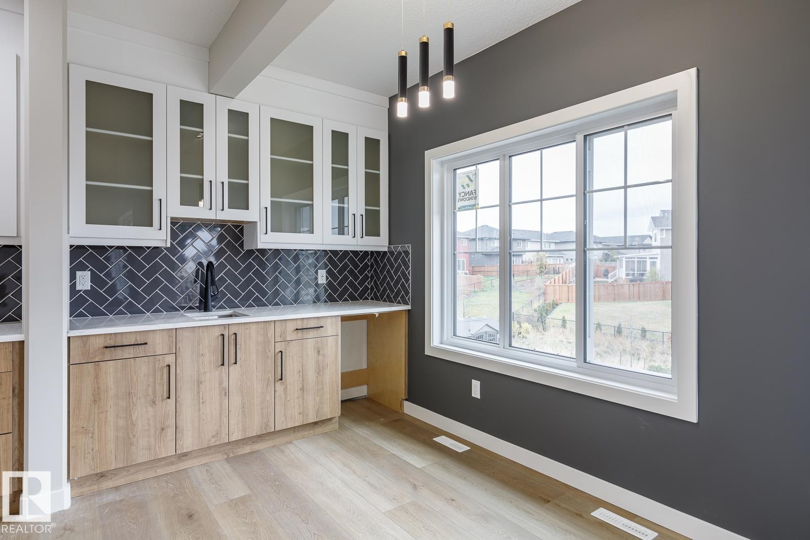 Kitchenette featuring white upper cabinetry with glass inserts, wood-finish lower cabinetry, a black herringbone tile backsplash, a sink with a matte black faucet, and light wood-finish flooring - 917 Elderberry Landing, Edmonton, AB - Indoor