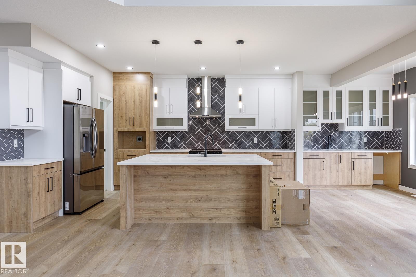 Spacious kitchen featuring an expansive island with a light-toned countertop, dual-tone cabinetry with wood-finish and white uppers, stainless steel appliances, a dark herringbone tile backsplash, and recessed lighting - 917 Elderberry Landing, Edmonton, AB - Indoor Photo Showing Kitchen