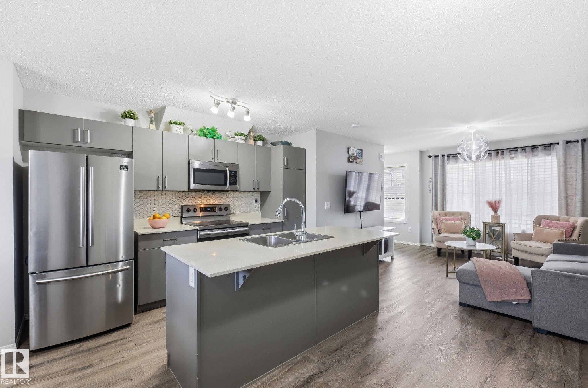The open concept living area features resilient plank flooring and a kitchen with stainless steel appliances, a white quartz countertop island, and gray cabinetry - 4636 175 Ave, Edmonton, AB - Indoor Photo Showing Kitchen With Double Sink